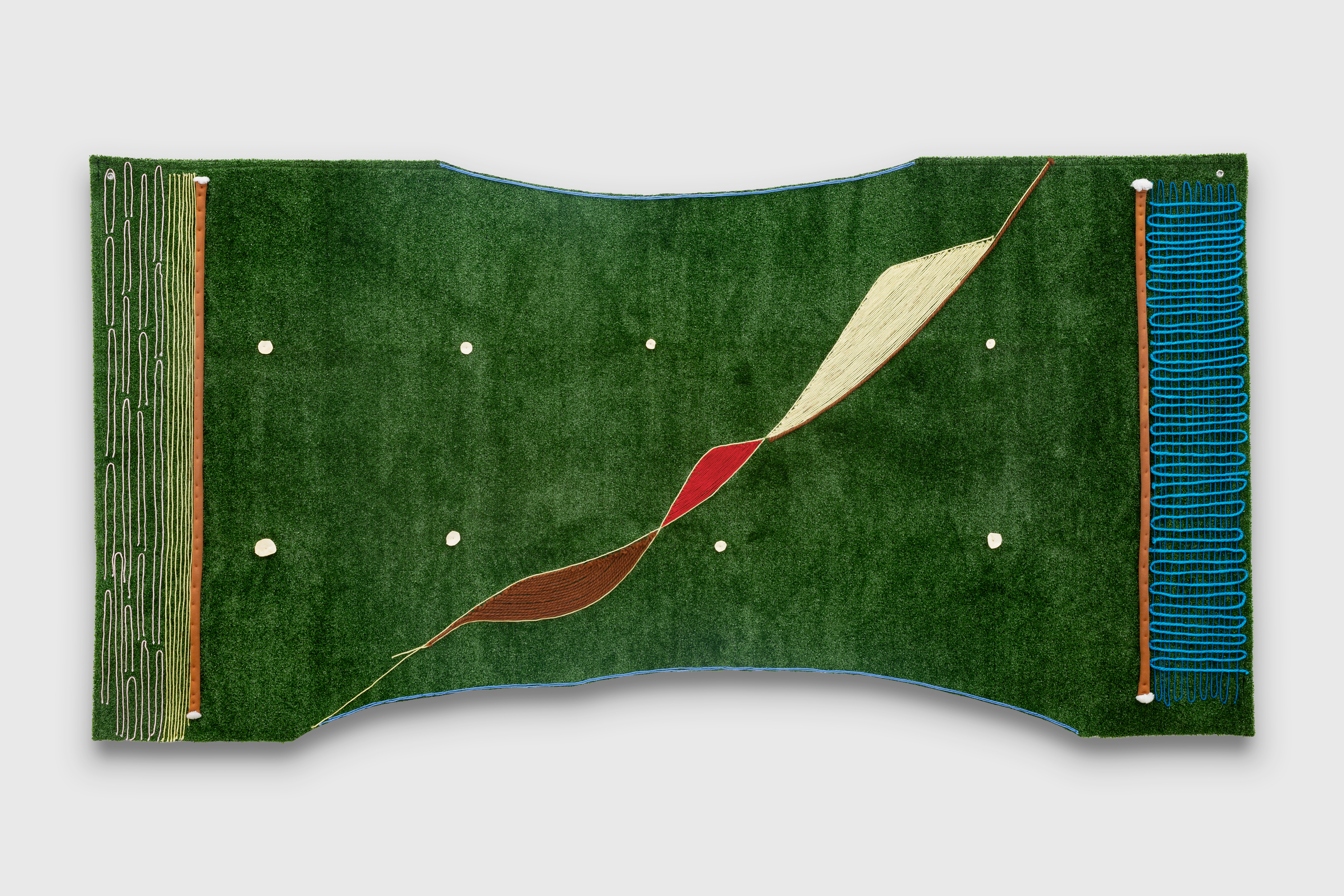 Close up of “Move In A Pattern” installed on a white wall. A rectangular shape in football-stadium green with two semicircular, inward curves in the center of the horizontal top and bottom edges, which are outlined with blue yarn. The top indentation encompasses about the entire center of the artwork. The bottom indentation covers about two thirds making the bottom edges to more prominent.  Beginning at the very left of the bottom indentation and running diagonal, reminiscent of a hammock that is twisted in the center, are three shapes that connect with one another. From bottom to top, they are brown yarn outlined in cream yarn, red year outlined in cream, and cream year outlined in red on one side only. The edges are decorated with yarn, buckskin, and artificial sinew, formed in various squiggly and/or linear patterns. Ten small white circular objects are spread out throughout the entirety of the work.