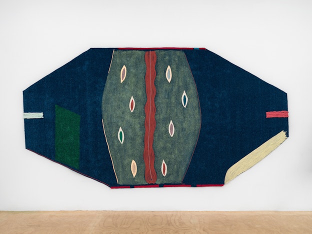 Close up of “Waking”, a dark blue, wide irregular octagon canvas on astroturf installed on a white wall. From the top edge of the hexagon to the bottom is an organic shape painted in a washed-denim blue, which has 8 pod-like shapes inside of it in pink, red, green, blue, or beige, with a white or blue lip. Additional shapes and borders to shapes are spread throughout the work in varying colors of yarn, buckskin, or paint on top of AstroTurf.