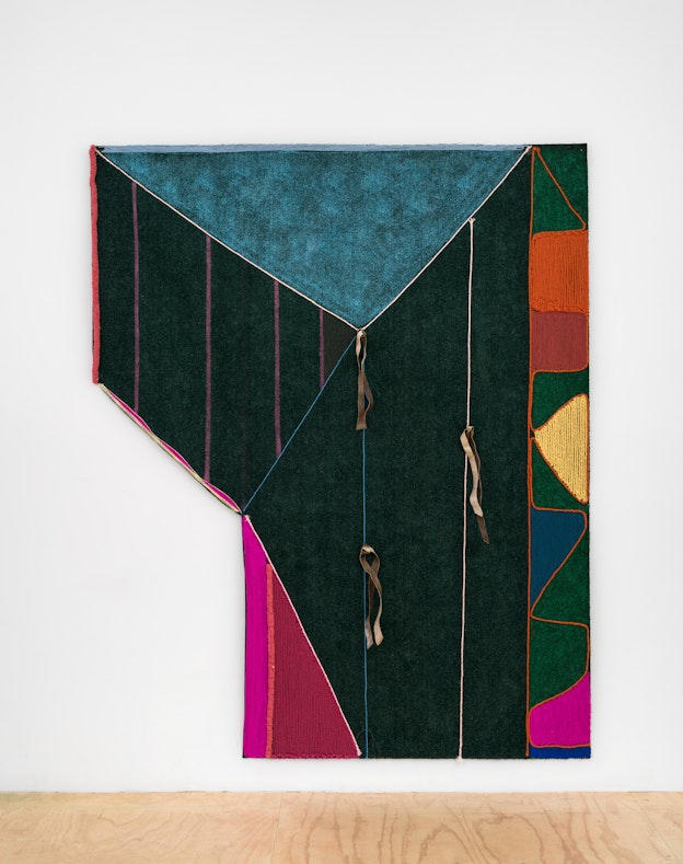 Close up of “Tracing the Memory”, a large AstroTurf artwork installed on a white gallery wall above a wooden floor. Asymmetrical geometric panels in deep green, teal, magenta, orange, red, and blue are stitched to one another with yarn and/or artificial sinew. Three ribbons made of buckskin hang off of the center of the piece.