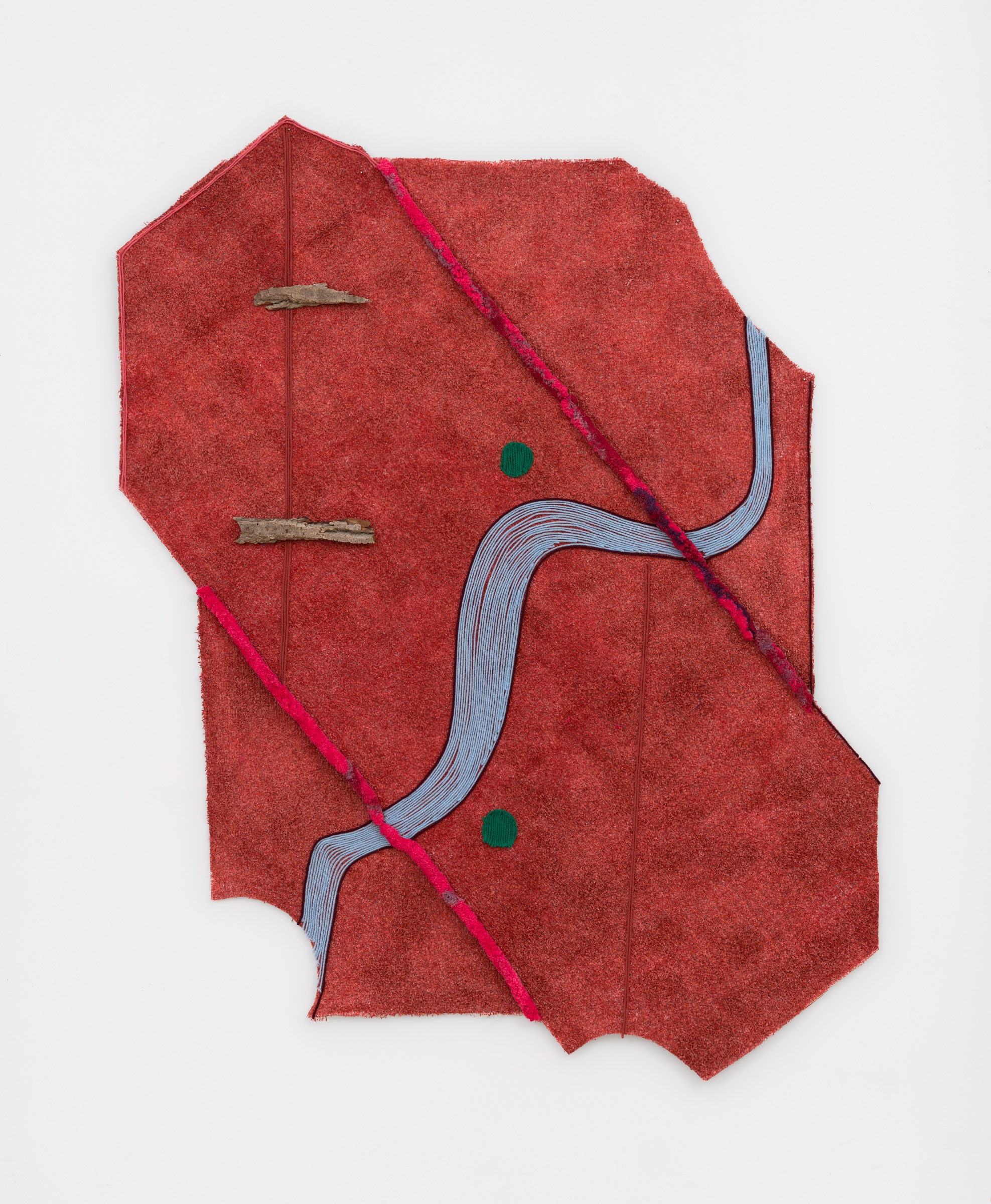 Close up of “Of This Time”, a large red irregular pentadecagon made of AstroTurf installed on a white gallery wall. Light blue yarn with a black border, reminiscent of a map-view of a river, is stitched into the astroturf, running across from the bottom left of the artwork to the top right corner. Above and below the center of the ‘river’ are two green circles made of yarn, also stitched into the AstroTurf. Two pieces of cottonwood lay horizontal in the top left corner. Finally, resembling pipe cleaners, two thick pieces of fuchsia yarn with indigo, violet, and red spots throughout, run diagonal and parallel to one another from left to right.