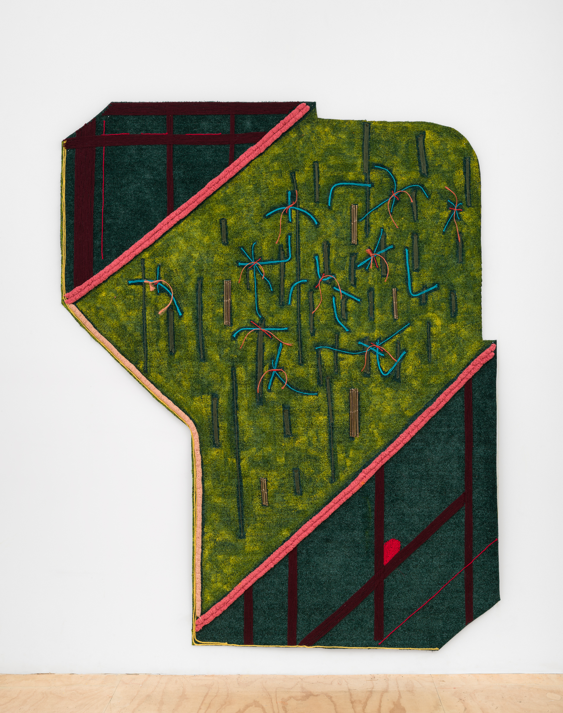 Close up of “Movements of The Land” a large organic 12-sided shape installed on a white gallery wall above a wooden floor. The shape’s body is predominantly a tennis court green, which has bright blue yarn stitched throughout in shapes that resemble roots. Red and orange yarn hangs off of the roots’ center in areas. There is gray yarn stitched throughout this section in vertical lines, as well as pieces of willow and hay bale rope. Bordering the tennis court green section with pink yarn, in the left and right corners of the artwork, are two irregular trapezoids made of AstroTurf painted in deep green with maroon trapezoids or rectangles stitched in with yarn, in addition to some red and yellow lines and a red shape that resembles a gem.
