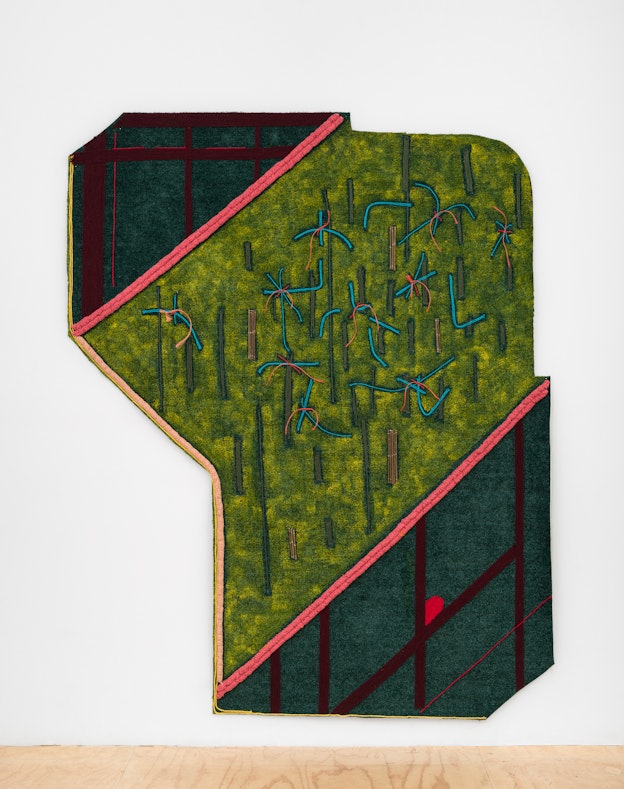 Close up of “Movements of The Land” a large organic 12-sided shape installed on a white gallery wall above a wooden floor. The shape’s body is predominantly a tennis court green, which has bright blue yarn stitched throughout in shapes that resemble roots. Red and orange yarn hangs off of the roots’ center in areas. There is gray yarn stitched throughout this section in vertical lines, as well as pieces of willow and hay bale rope. Bordering the tennis court green section with pink yarn, in the left and right corners of the artwork, are two irregular trapezoids made of AstroTurf painted in deep green with maroon trapezoids or rectangles stitched in with yarn, in addition to some red and yellow lines and a red shape that resembles a gem.