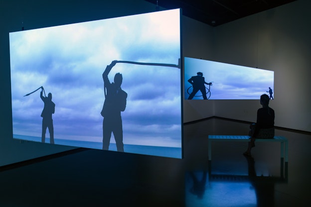 Installation view of “Gas Men,” a video work on two screens that hang from the ceiling in a gallery space. Projected on the screens are two different time stamps of the film. On the screen to the left, which is the larger screen, is a still that includes a silhouette of two people in suits swinging gas pumps over their head, in front of an ocean’s horizon and a blue sky with low puffy clouds. On the screen to the right are the silhouettes of two people in suits, who appear to be fighting each other with the gas pumps, against a blue sky with puffy clouds. The screen on the right reflects onto the grey floor, as well as a small white bench, where someone is seated and looking at that screen. Behind the projection screens are two white gallery walls.