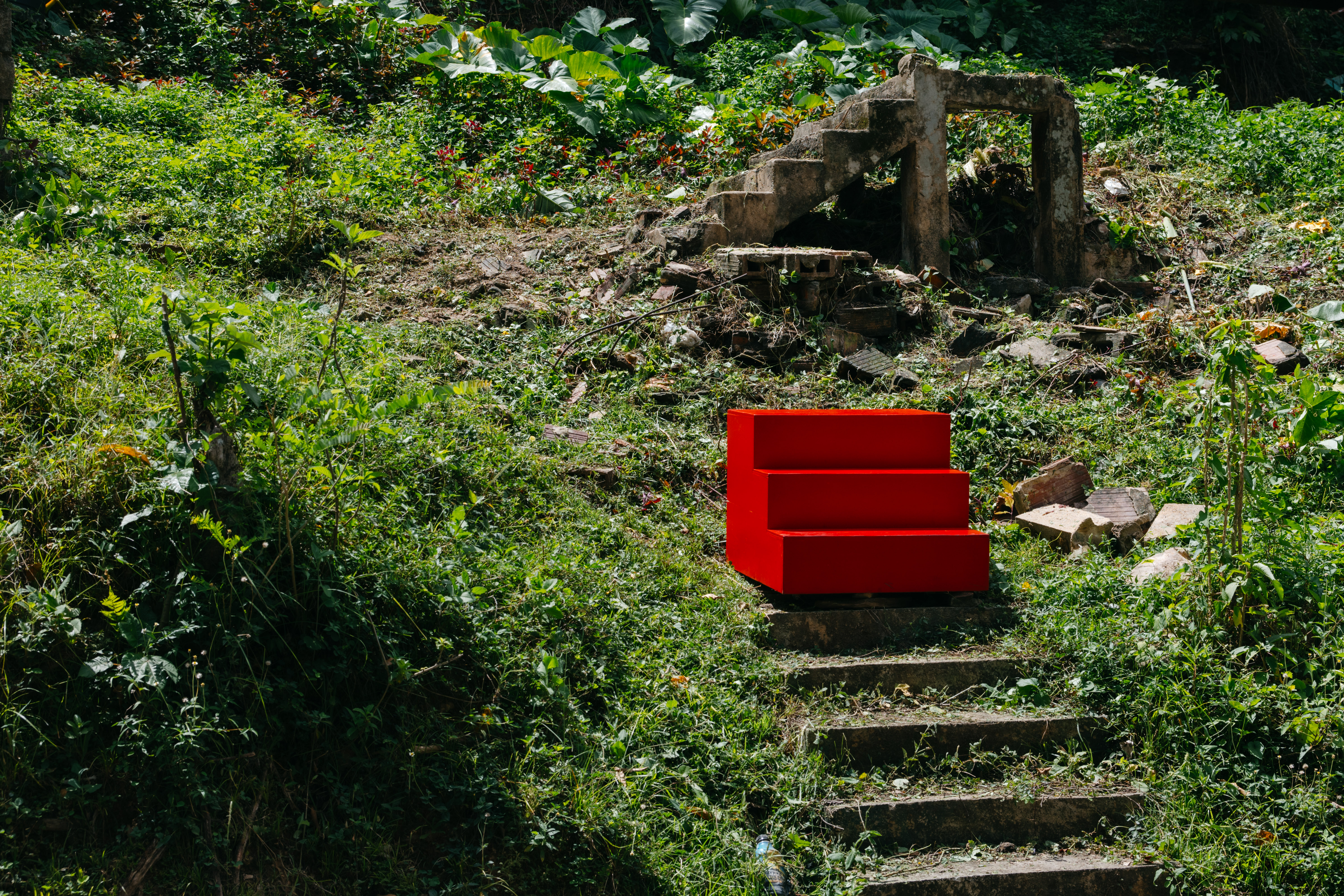 Installation view of “Home/Portal,” a red three-step stairway, which is placed on top of a stone staircase that runs through a sun-lit hill in a vibrant park or garden. There is grass all along the stone staircase and growing out of it. Lush green plants and little plants with red flowers are scattered throughout the hill. Disconnected from the stone staircase are five more steps made out of stone. The steps that would have connected the two are in ruins, with their stones dispersed throughout the grass.