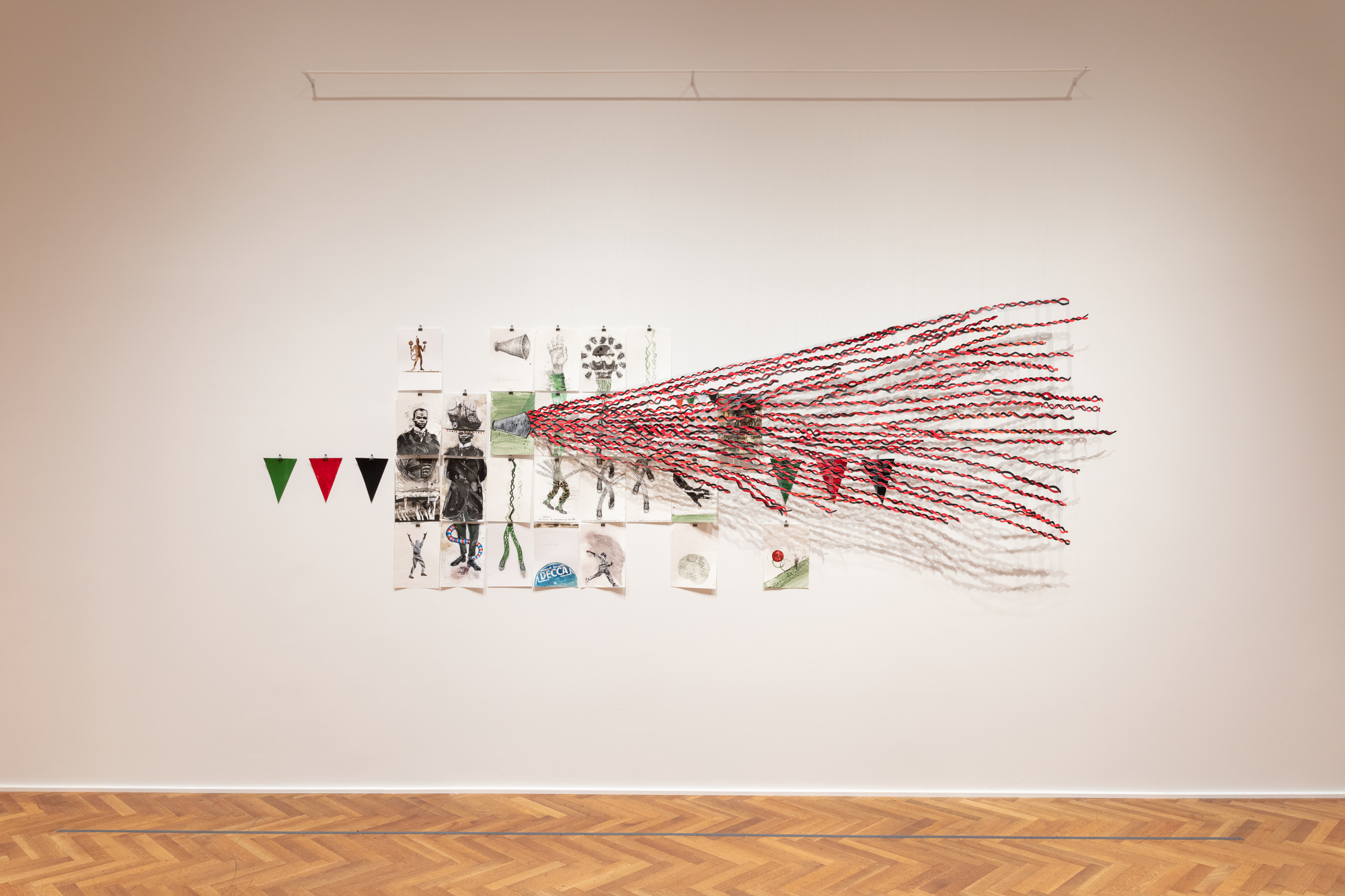 Installation view of “Stepping,” against a white wall above a  herringbone-patterned wooden floor. An uneven grid of 28 pieces  of paper with drawings on them are individually hung on the wall  with binder clips. To the left and right side of the grid are three  pennant flags in green, red, and white, also separately attached  to the wall with binder clips. Drawings are of men, hands, legs,  figures, snakes, and amplification devices, among other objects,  in earthy tones and red. Several of the drawings are realized on  two pieces of paper, while the rest only make use of one. In the  center of the grid is an amplification device that extends outward  beyond the paper through braided red and black cords. The cords  vary slightly in length and spacing, which creates a dynamic,  wave-like form that casts layered shadows on the wall behind it.