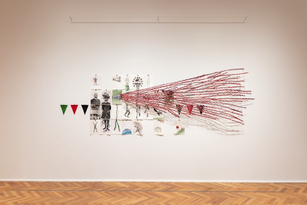 Installation view of “Stepping,” against a white wall above a  herringbone-patterned wooden floor. An uneven grid of 28 pieces  of paper with drawings on them are individually hung on the wall  with binder clips. To the left and right side of the grid are three  pennant flags in green, red, and white, also separately attached  to the wall with binder clips. Drawings are of men, hands, legs,  figures, snakes, and amplification devices, among other objects,  in earthy tones and red. Several of the drawings are realized on  two pieces of paper, while the rest only make use of one. In the  center of the grid is an amplification device that extends outward  beyond the paper through braided red and black cords. The cords  vary slightly in length and spacing, which creates a dynamic,  wave-like form that casts layered shadows on the wall behind it.