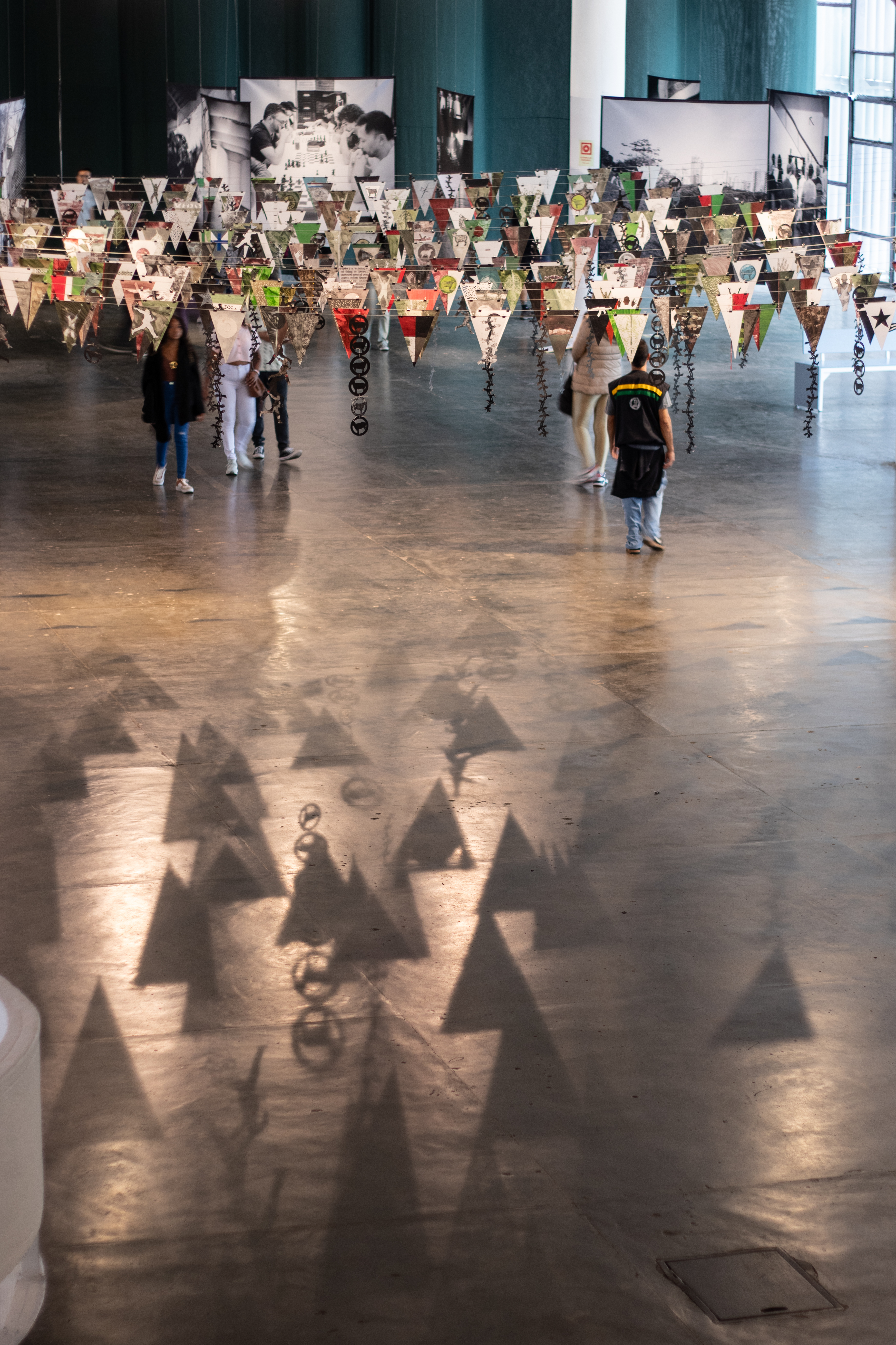 Installation view of “After the Appeal Will Come the Next Delivery,” which is many rows of pennant flags interspersed with one another. The flags have varying drawings and colors on them, mainly using earth tones, as well as bright red and green. Symbols made of black paper hang with the flags and patrons of the biennial walk under the flags, which have shadows reflecting onto the grey stone floor. Black and white digital prints are installed behind the work “Stepping.”