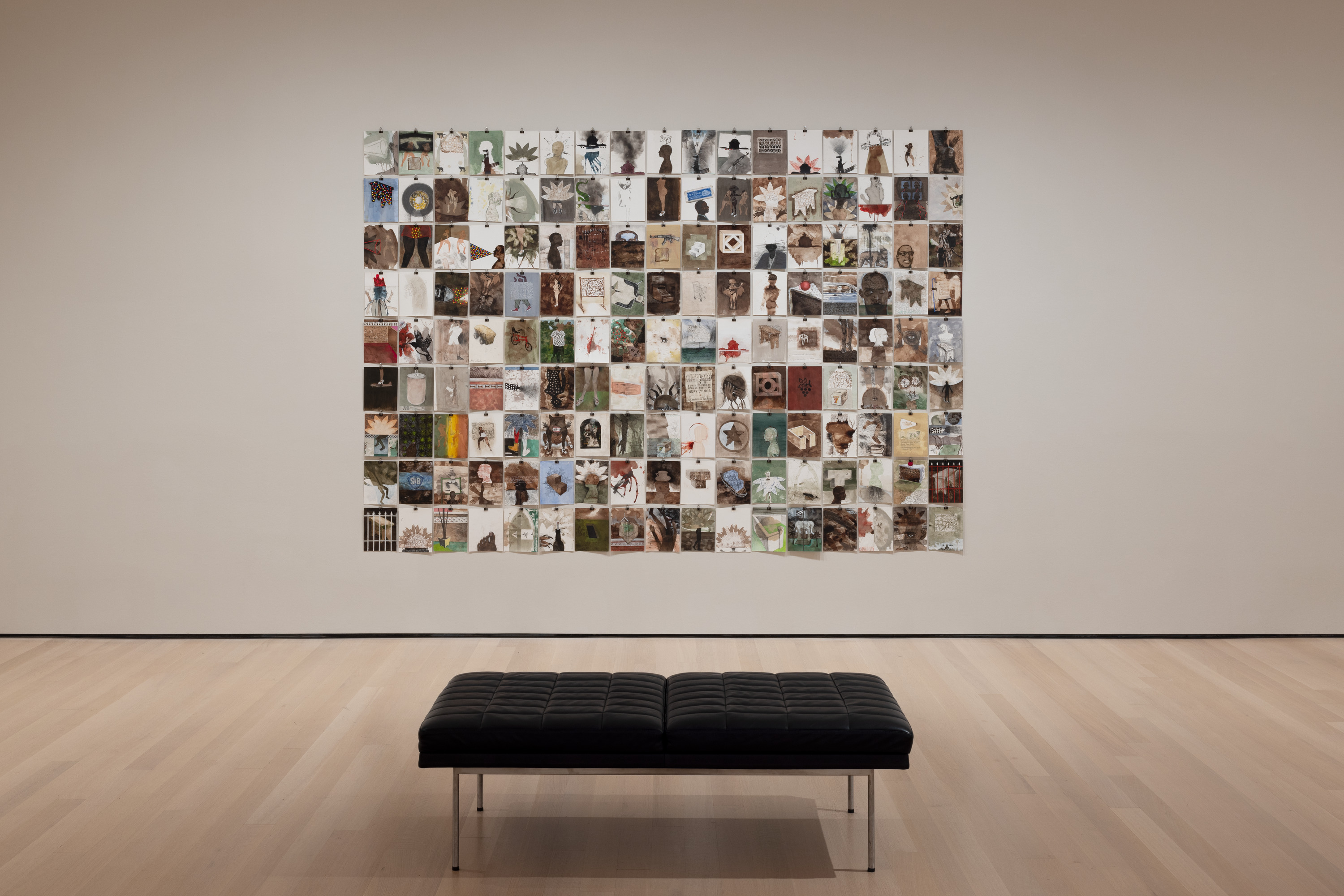 Installation view of “Tropical Night,” a grid of drawings on 153 individual rectangular pieces of paper, each of which is attached to a white museum wall with a binder clip. The drawings all vary in images and colors, with recurring themes of hands, feet, plants, stars, guns, and infrastructure, in earth tones and reds. In front of the artwork, on the wooden floor, is a bench with a black cushion and silver legs.