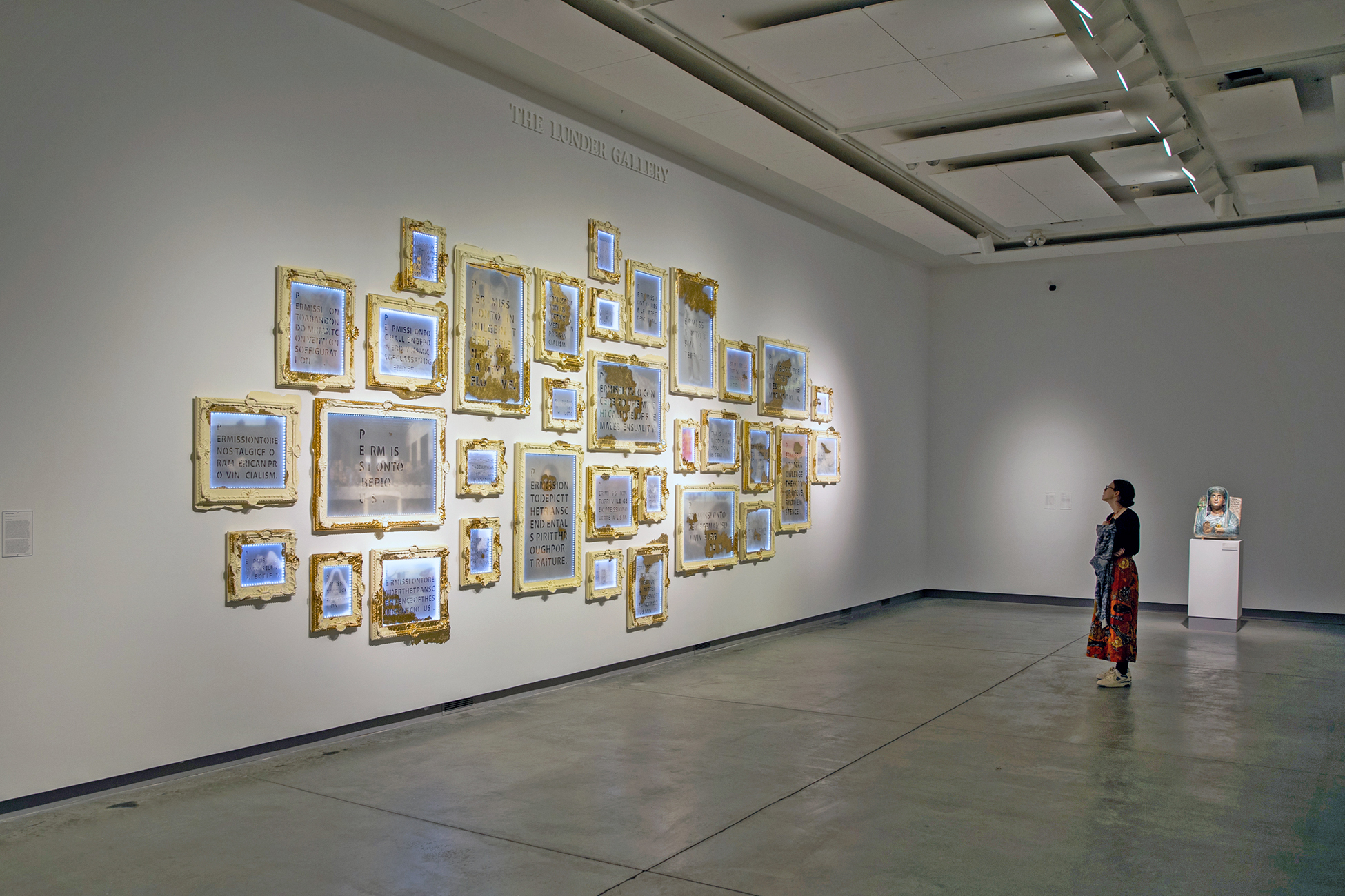 Installation view of “Soft Power. Hard Margins.” 32 thematic artworks are placed  close to one another on a white gallery wall. Each individual piece includes a light  blue or grey mixed-media paper with text, which is illegible due to the distance the  photo was taken with, and is framed by gold and/or cream acrylic, cast resin, wood,  and metal leafs. White LEDS also border the paper or the rabbet of the frame. A  patron stands on grey stone floor tiles, engaging with Morgan’s work. Behind them  and against the neighboring wall is a white pedestal stand featuring “May the  obdurate foe not be in good health,” a palmyrene funerary bust by Michael Rakowitz.