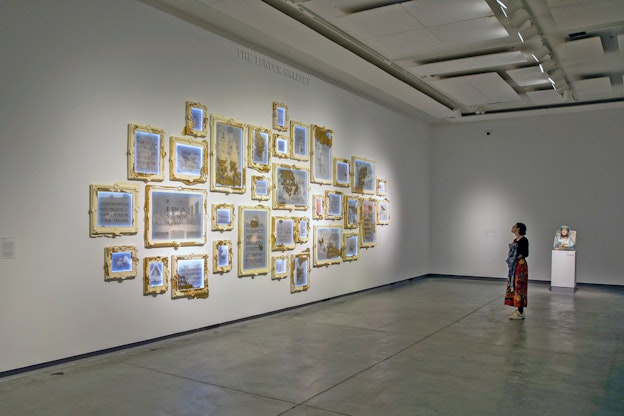 Installation view of “Soft Power. Hard Margins.” 32 thematic artworks are placed  close to one another on a white gallery wall. Each individual piece includes a light  blue or grey mixed-media paper with text, which is illegible due to the distance the  photo was taken with, and is framed by gold and/or cream acrylic, cast resin, wood,  and metal leafs. White LEDS also border the paper or the rabbet of the frame. A  patron stands on grey stone floor tiles, engaging with Morgan’s work. Behind them  and against the neighboring wall is a white pedestal stand featuring “May the  obdurate foe not be in good health,” a palmyrene funerary bust by Michael Rakowitz.