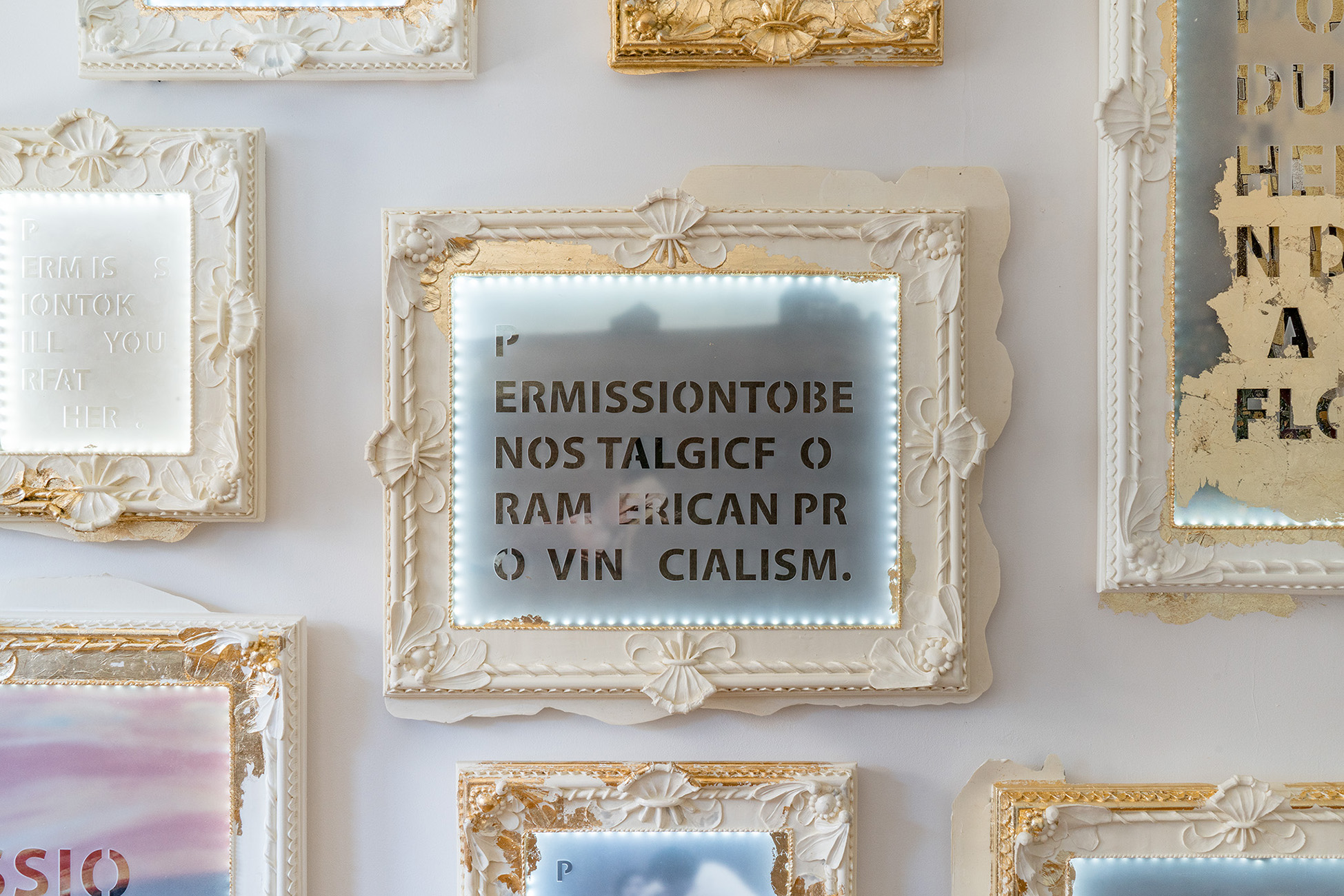 Close up shot of a light blue mixed media paper that reads “P ERMISSIONTOBE NOS TALGIC  F () RAM ERICAN PR O VIN   CIALISM,” which is one work in the “Soft Power. Hard Margins.”  Installation. It is framed by cream acrylic, cast resin, wood, and metal leafs. White LEDS also  border the paper, or the rabbet of the frame. Additional frames with mixed media paper, all of  which are cut off in part, encircle this work.