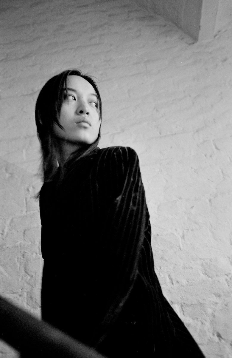 Black and white portrait of Pan Daijing. Pan stands at an angle dressed in all black against a white textured wall. She looks off to the distance.
