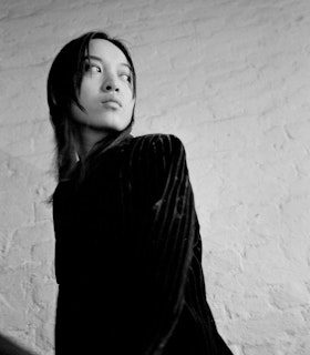 Black and white portrait of Pan Daijing. Pan stands at an angle dressed in all black against a white textured wall. She looks off to the distance.