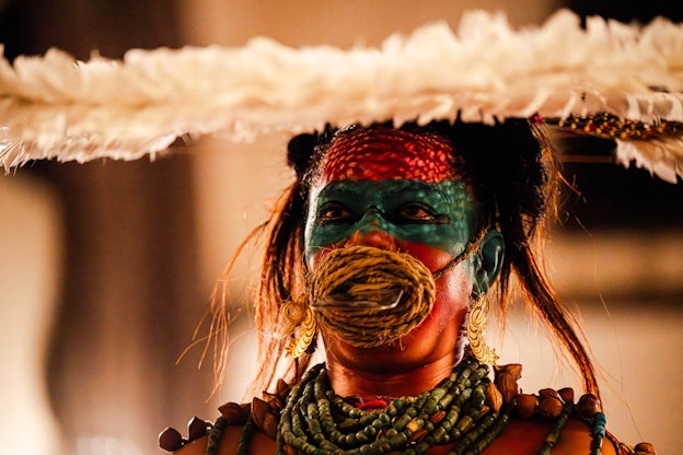 Mid-performance close up of Lukas Avendaño. Avendaño is looking up at the front of her feather  headdress as a yellow rope is coiled around her mouth and looped around her head, just over her ears, which  are decorated with long gold earrings. Inside the center of the rope coiled at her mouth is a flat silver object,  which juts outwards. Her face is painted red and blue. A beaded turquoise necklace is wrapped around  Avendaño's neck in multiple layers. The image is cut just after her shoulders, where she wears ayoyotl.