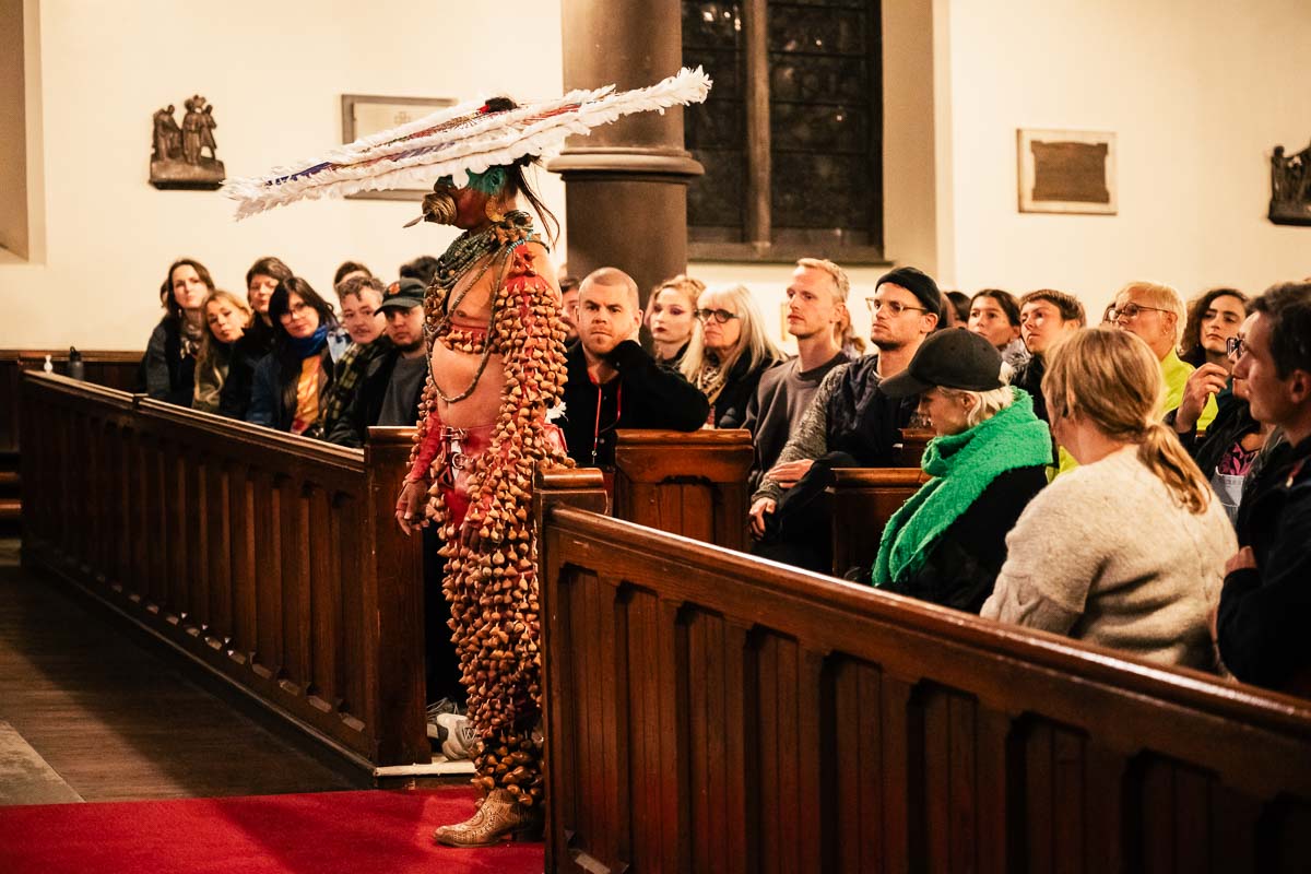 Side view of Lukas Avendaño mid performance. Wearing a costume made of ayoyotl and a large feather  headdress, Avendaño stands with her back to the audience on a red runner rug between the front church  pews. The audience is seated throughout the pews.