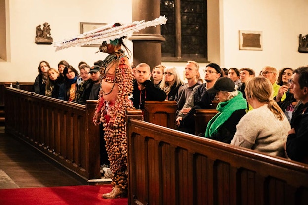Side view of Lukas Avendaño mid performance. Wearing a costume made of ayoyotl and a large feather  headdress, Avendaño stands with her back to the audience on a red runner rug between the front church  pews. The audience is seated throughout the pews.