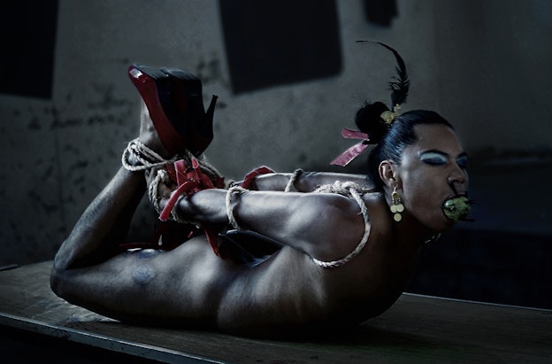 Lukas Avendaño mid-performance in a dimly lit room with paint chipped off the walls. Avendaño is tied  up, with her stomach against a wooden table, wearing only red high heals. Her shoulders, elbows, hands,  and ankles are bound together with a beige rope, while her forearm and wrists are tied with a makeshift  red rope. Avendaño’s eyes are closed and her mouth gagged with a green ball reminiscent of a prickly  pear cactus pierced with black spikes. Gold earrings dangle from her ear and her hair is decorated with  a black feather, pink ribbon, and yellow accessory. 
