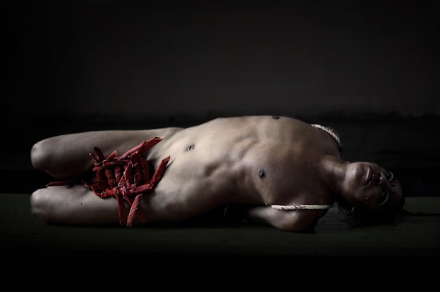 Lukas Avendaño mid-performance. Avendaño is laying down to the side and undressed as her arms and  feet are bound behind her with a beige rope. An assortment of tapered red cloths are knotted together  throughout her genital region and between her legs. Avendaño is illuminated by a soft spotlight and the  rest of the room is dark.