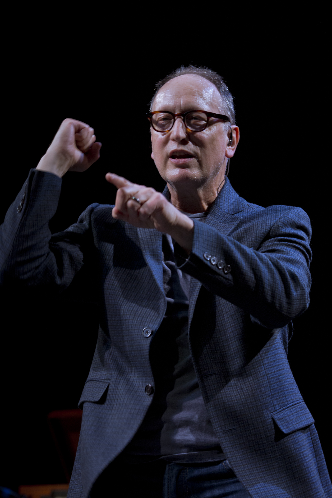 Paul Lazar mid performance in a pitch black room. Lazar opens his  mouth while pointing with his right index finger, and making a fist with  his left hand. He’s wearing an unbuttoned blue and black checkered  suit, a blue shirt underneath, and blue jeans. The photo is cropped  right below his waist. 