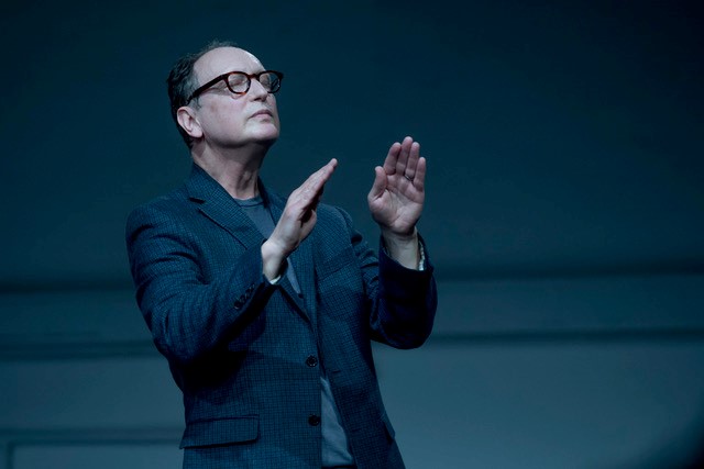 Paul Lazar mid performance in a blue room. Lazar’s eyes are closed as  he appears to be conducting with his hands. He is wearing a blue and  black checkered suit jacket, which is buttoned only in the center, with  a blue shirt underneath. The photo is cropped at his waist. 