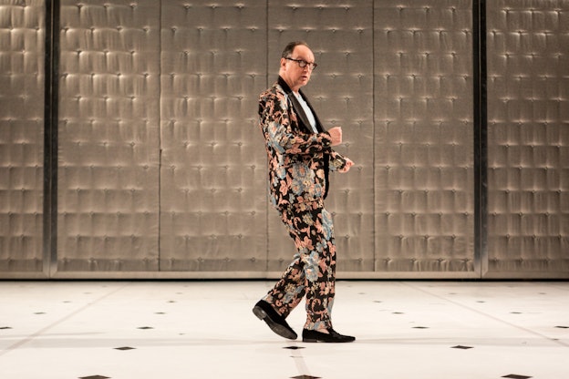 Paul Lazar mid performance in front of a tufted beige wall. He’s wearing  a pink, blue, and black floral patterned suit with a white shirt underneath  the jacket and black dress shoes. Lazar's hands are in fists and he has  one foot in front of the other, with his back leg just off the ground. The  flooring is cream, garnished with straight lines of black diamonds.