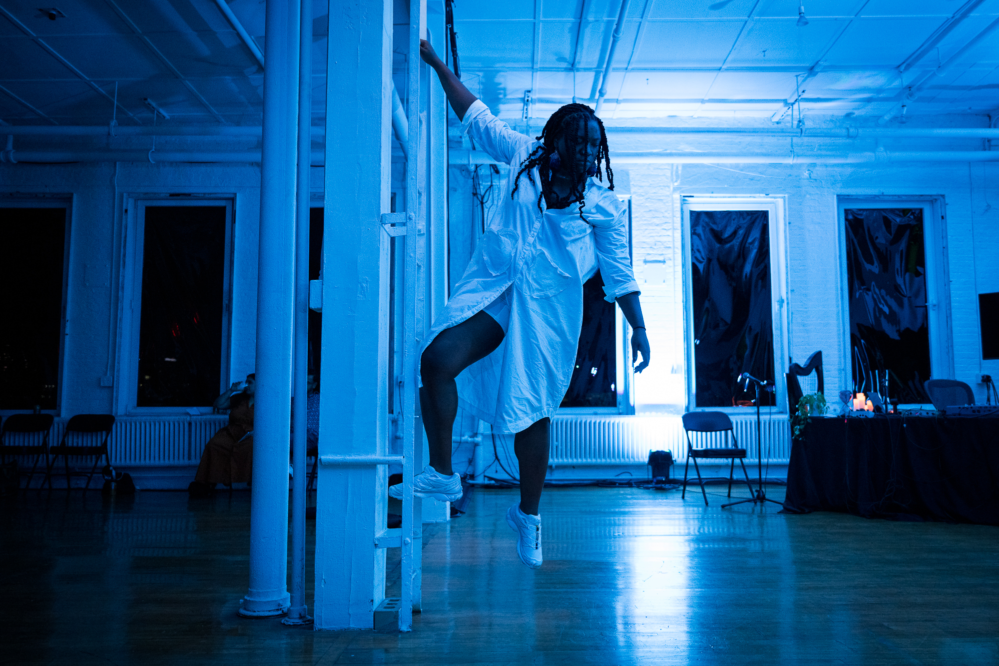 Ogemdi Ude is climbing a ladder that is built into the floor at The Kitchen.  She is looking down with one foot and hand in the air. The light blue lighting  in the room reflects on Ude’s white clothing and shoes, the ladder, the pipes,  and the wall. Behind Ude, in the corners of the room, are a table with a black  table cloth that features a small green plant and several candles glowing  with a pink shade, a harp, several chairs spread throughout the room,  lighting, and six windows with blackout curtains.