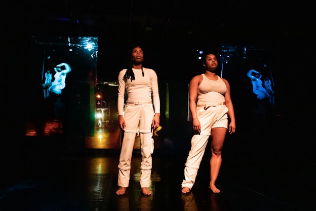Runako Campbell and Selah Hampton stand with their hands to their sides, looking  away and above the camera with lighting pointed towards them. They are both  wearing all white and barefoot, between two projector screens displaying a previous  moment of the performance in flipped views. The projector display to the left is tinted  light blue, while the one to the right is a darker shade of blue. The ceiling is without  lighting but the back of the room features bright yellow, red, and green lights. 