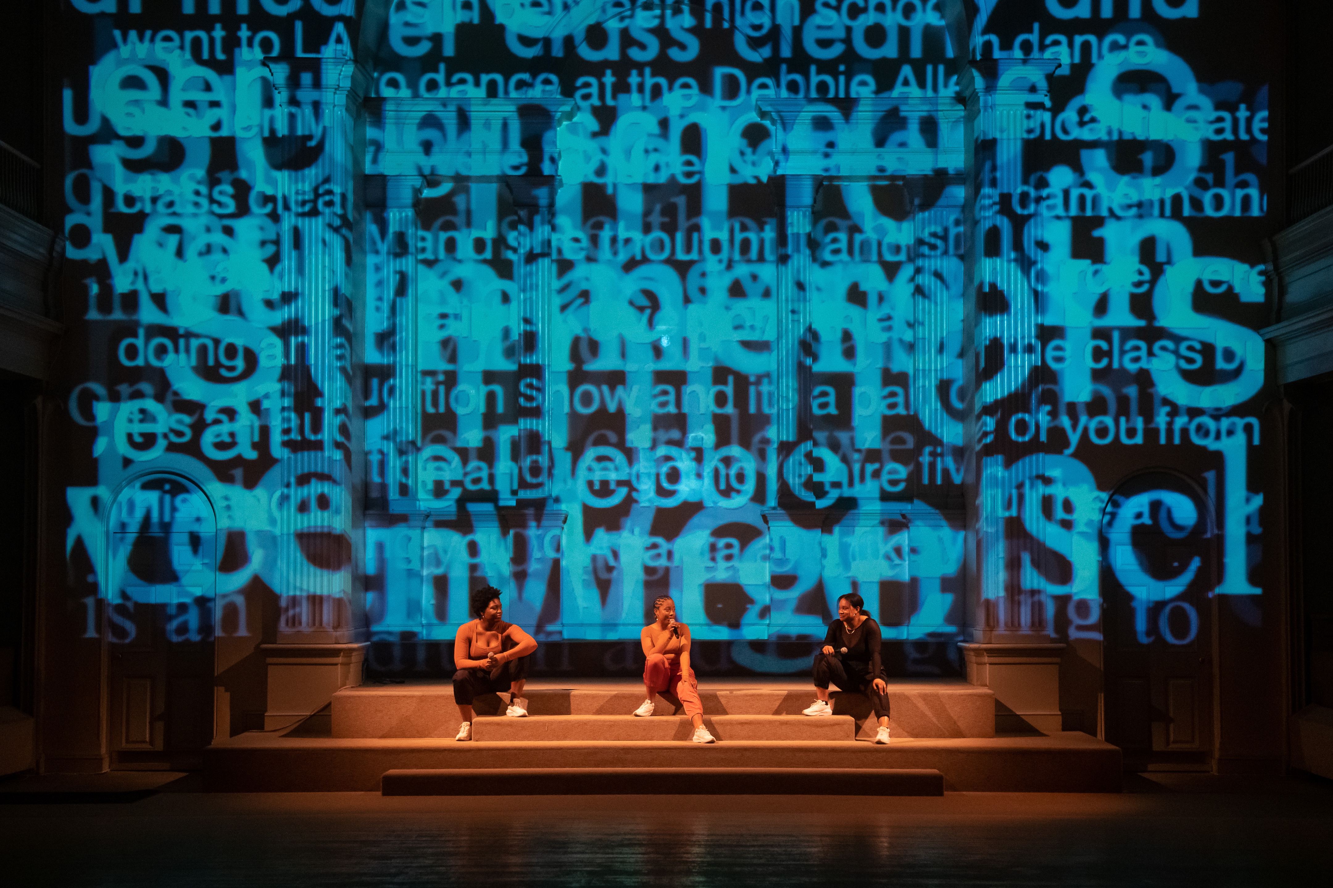 Ogemdi Ude, Symara Sarai, and Selah Hampton sitting on carpeted steps and talking to  each other in a performance space. Each of them is holding a microphone and wearing  white shoes, with one foot resting on a step above the other. Projected on the wall is  light blue text that layers over itself. Some of the words that can be seen include  “went to LA to dance at the Debbie”, “dance”, “is an”, “show and its a”, “to, “of you  from”, “doing”, and “class”.
