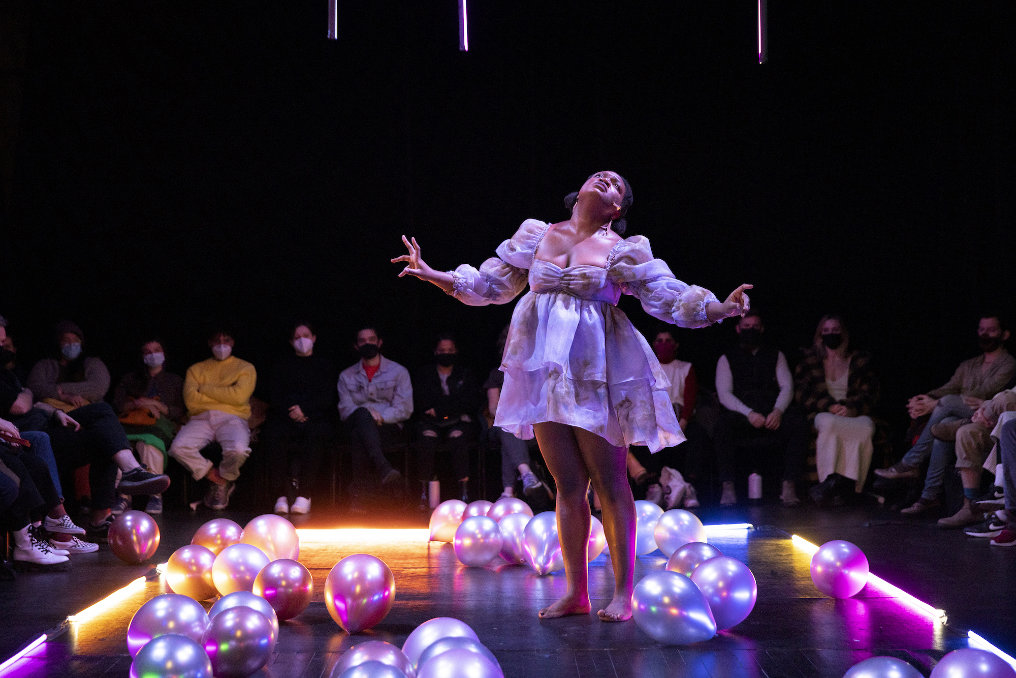 Ogemdi Ude stands in the middle of the room with her hands sustained in the air.  She is surrounded by shiny balloons spread throughout the floor, which reflect the  white lighting coming from above, as well as the yellow, pink, and blue lighting that  encloses the dance floor. Ude is wearing a flowy baby-doll long sleeve dress, which  matches the colors of the balloons. Audience members are behind her and to her  sides, gazing towards her. Everyone in the audience is wearing a mask.