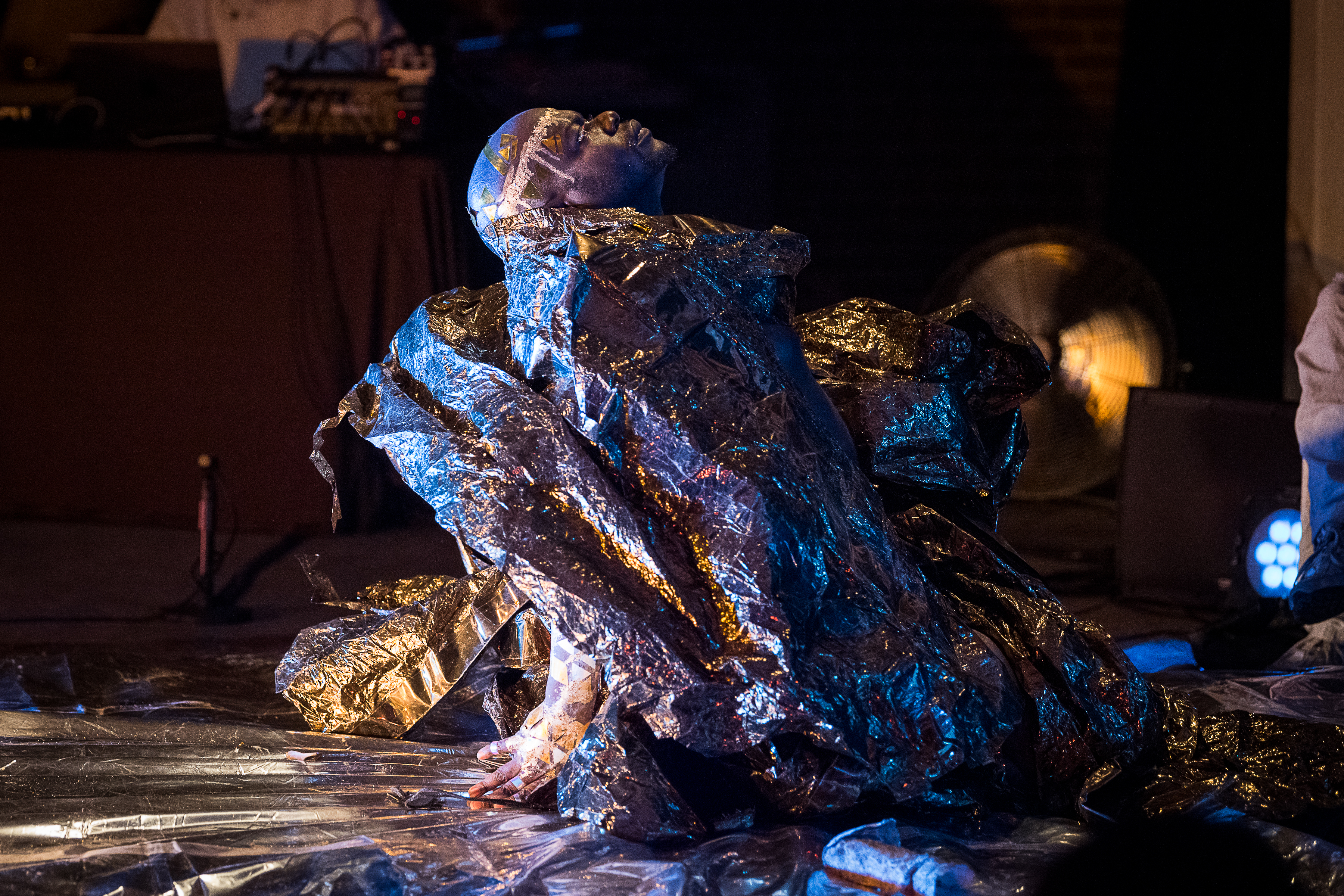jaamil olawale kosoko sits on their knees, looking up, with their hands behind their back. Spotlighted, they are wearing a shiny silver costume. The floor is wrapped in a plastic material.