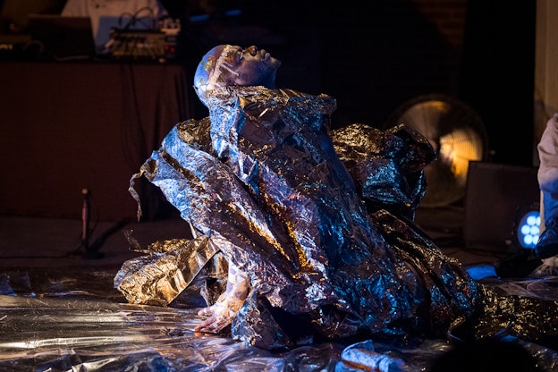 jaamil olawale kosoko sits on their knees, looking up, with their hands behind their back. Spotlighted, they are wearing a shiny silver costume. The floor is wrapped in a plastic material.