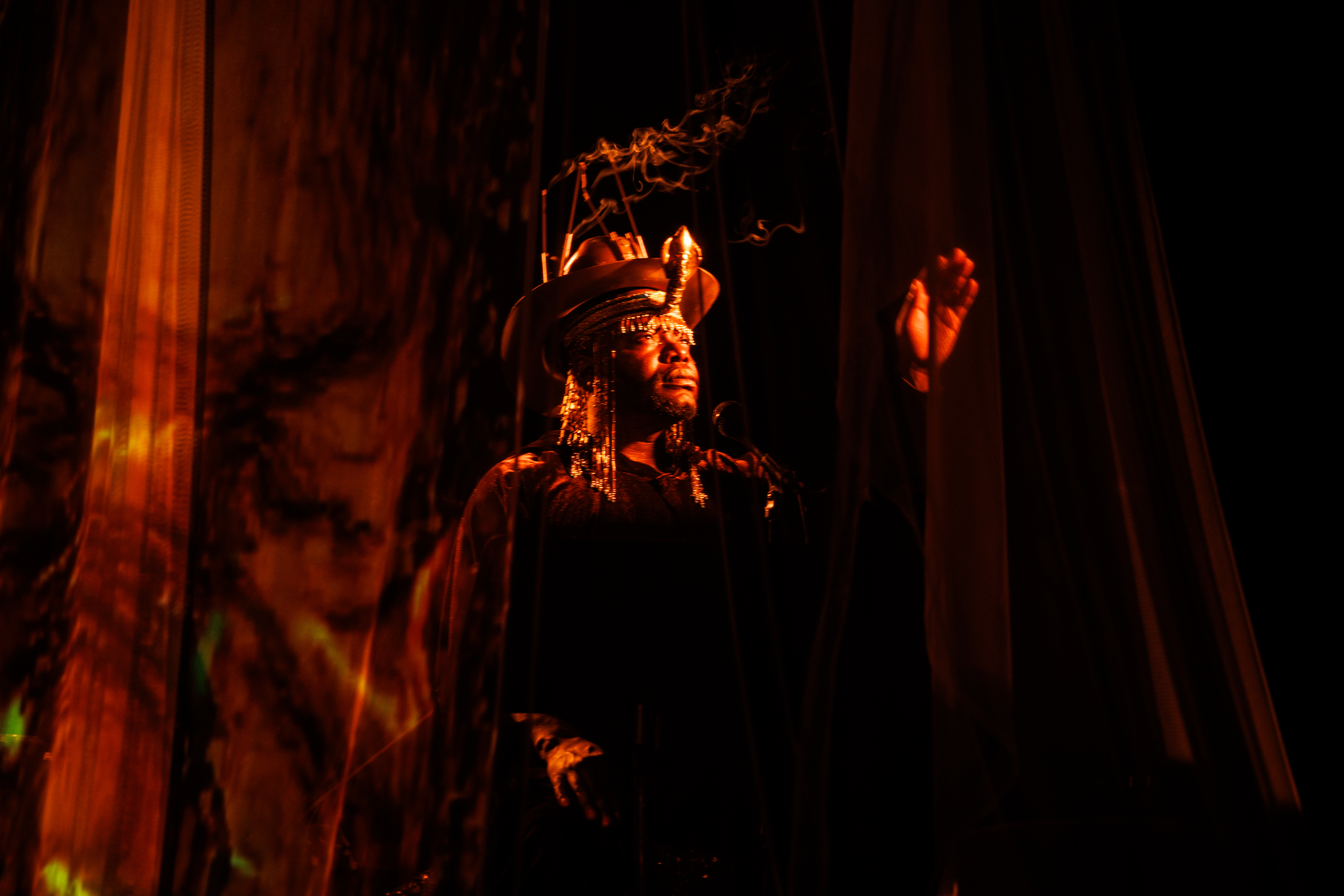 jaamil olawale kosoko performing in red and orange lighting in between transparent curtains. They have one hand up, with their palm facing the audience. kosoko is wearing a hat, on which sits a a cone shaped structure emitting smoke.