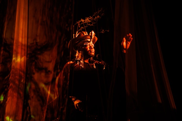 jaamil olawale kosoko performing in red and orange lighting in between transparent curtains. They have one hand up, with their palm facing the audience. kosoko is wearing a hat, on which sits a a cone shaped structure emitting smoke.