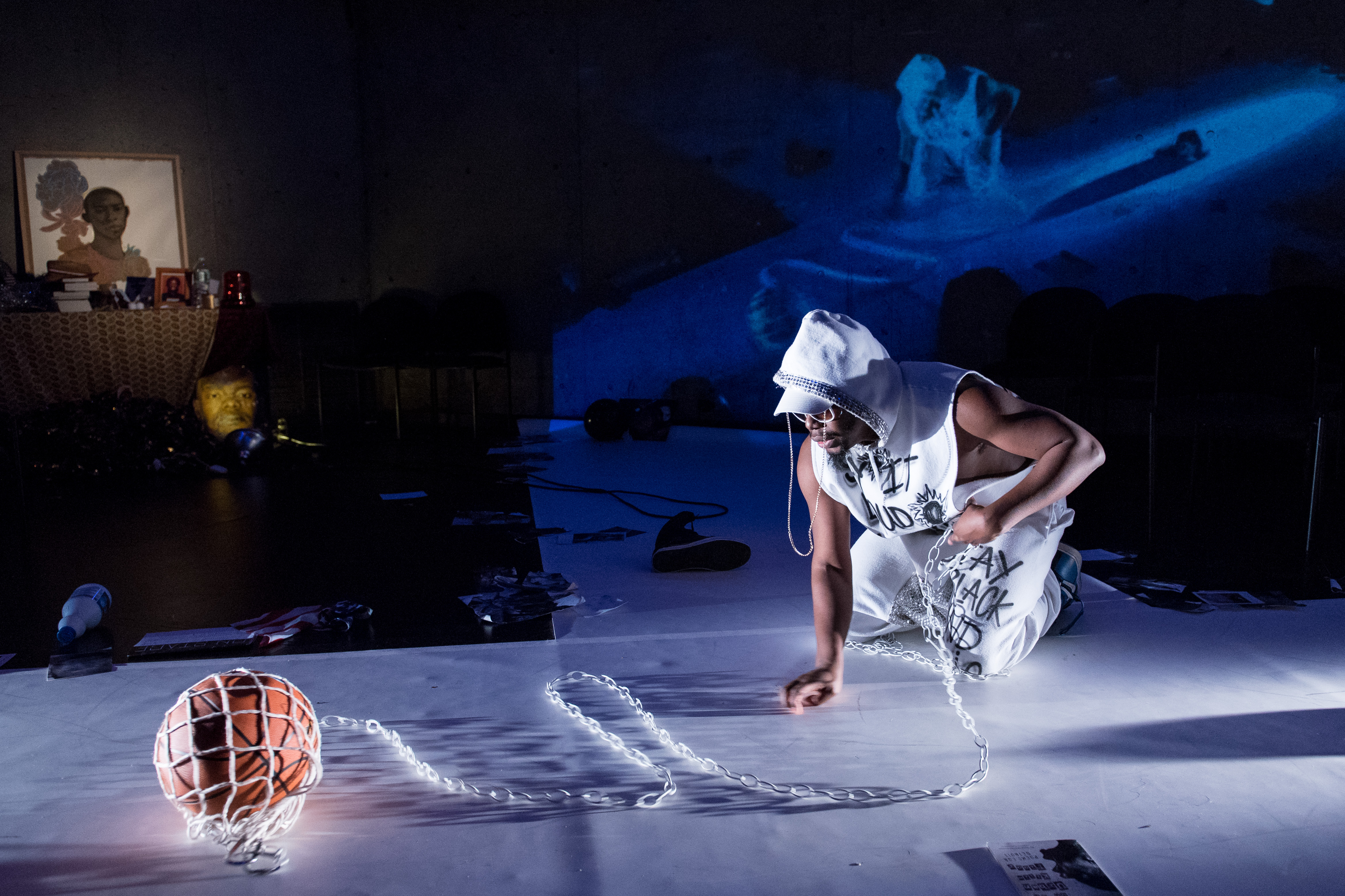 jaamil olawale kosoko kneeling on a white runway while holding a white chain connected to a basketball covered in a white mesh bag. They wear a white sweatsuit with the hood up, hat, and sunglasses. kosoko is projected on the wall behind themself in a blue hue. There is a shrine to dead Black people in the left hand corner, and the writings of Black  intellectuals spread throughout the room on the floor. Also on the floor is an American flag, a hightop shoe, a rope, and a bottle.