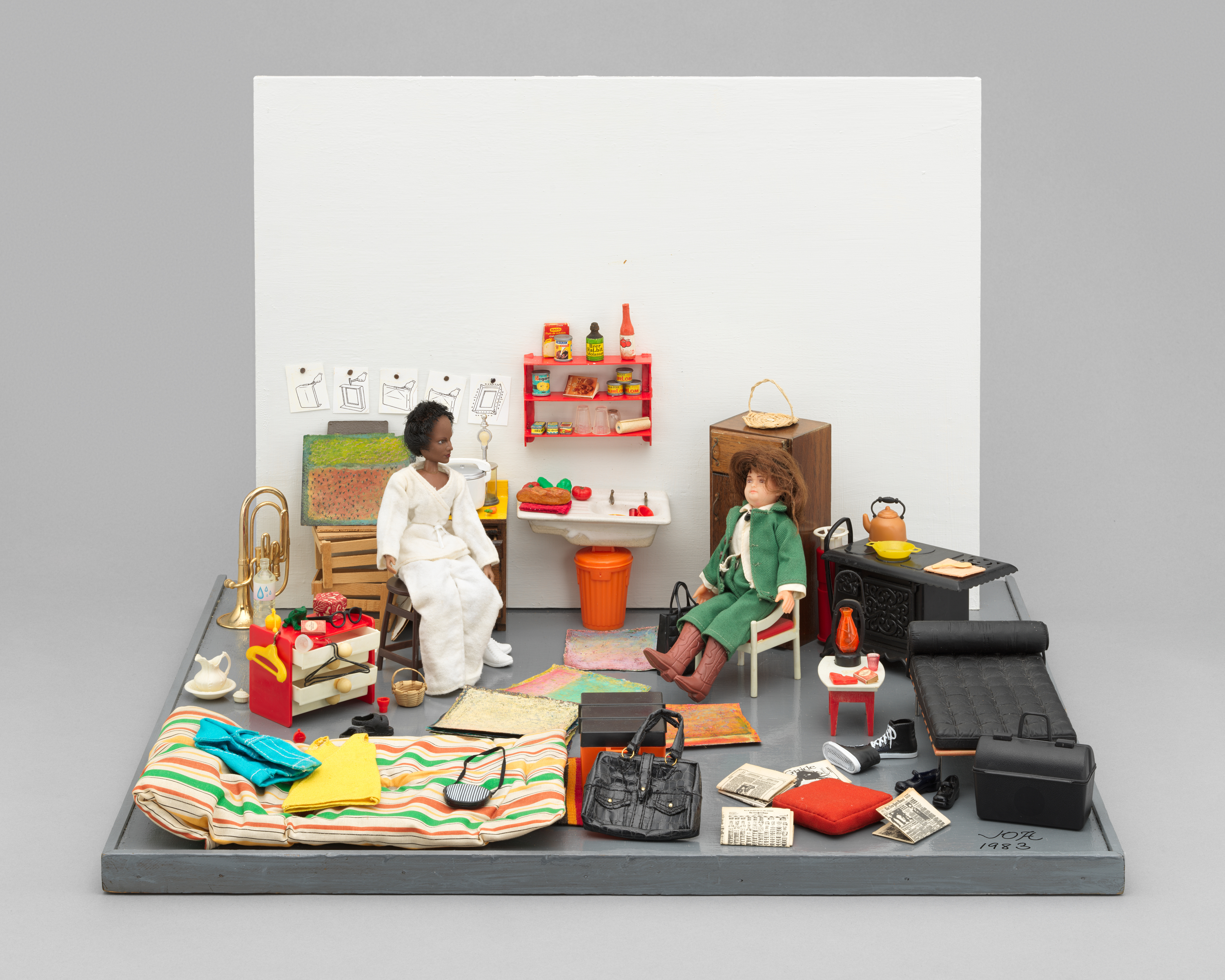 Installation view of “The Studio Visit,” a diorama sculpture of two dolls seated  in a small room cluttered with household items. The doll to the left is wearing  a white robe, looking directly at the doll to the right, who is wearing a green  coat over a white shirt, green pants, and brown cowboy boots, and looking  past her. In between and behind the dolls, in the center of the room, is a  white sink, orange trash can, and red shelf, which is fixed onto the diorama’s  white wall. Both the sink and the shelf have food and kitchen items on them.  The gray floor is scattered with rugs, bags, shoes, and newspapers, as well  as a trumpet, furniture, and personal objects. Notably, the objects on the left  side of the room make use of bright colors, while the right side of the room  includes predominately black items. Artwork is pinned or taped to the wall  on the left side of the room. The artist’s signature and date, “JOR 1983” is  handwritten in the bottom left corner of the room. The sculpture is against  gray wall, which is significantly lighter than the diorama’s gray floor.