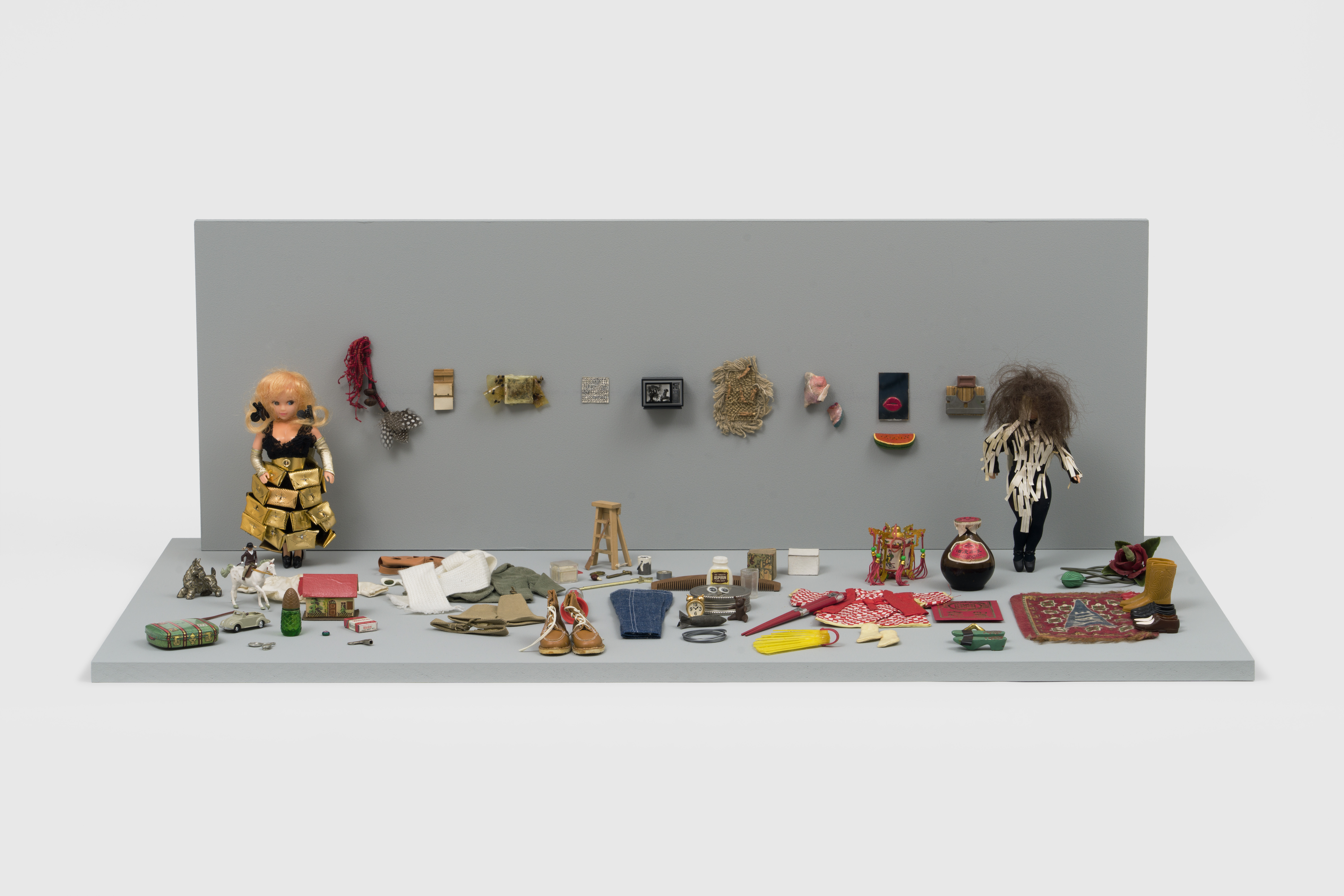 Installation view of “The Male Art Dependent,” a diorama sculpture of two dolls  that are on opposite ends of a room with objects scattered throughout. The doll  to the left wears a black lace dress with a shiny gold belt and a skirt of shiny  gold pocketbooks stapled onto lace, bronze arm warmers, two black hair  clips, and black shoes. The doll to the right wears a black unitard that is  unevenly decorated with off-white strips of paper. In between the two dolls,  on the grey wall, is a line of objects. The grey floor is filled with items such  as shoes, a rug, a vase, clothing, a ladder, a comb, a clock, measuring spoons,  a buggy car, sunglasses, a unicorn with a doll riding it, a pack of Winston  cigarettes, among other objects. The diorama is installed against a white wall.
