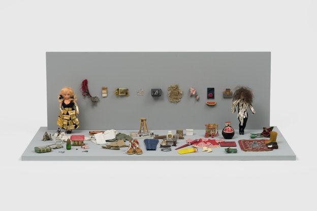 Installation view of “The Male Art Dependent,” a diorama sculpture of two dolls  that are on opposite ends of a room with objects scattered throughout. The doll  to the left wears a black lace dress with a shiny gold belt and a skirt of shiny  gold pocketbooks stapled onto lace, bronze arm warmers, two black hair  clips, and black shoes. The doll to the right wears a black unitard that is  unevenly decorated with off-white strips of paper. In between the two dolls,  on the grey wall, is a line of objects. The grey floor is filled with items such  as shoes, a rug, a vase, clothing, a ladder, a comb, a clock, measuring spoons,  a buggy car, sunglasses, a unicorn with a doll riding it, a pack of Winston  cigarettes, among other objects. The diorama is installed against a white wall.