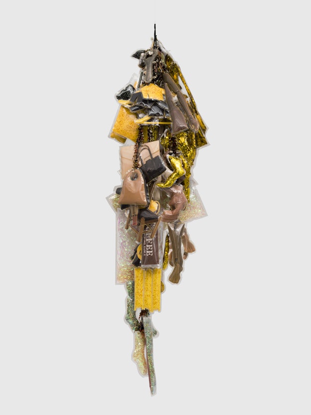 “Ritual (Self-Portrait),” a vertically hanging sculptural collage against a white  wall. The sculpture is made of dangling yellow, brown, gold, black, and white  objects, such as small handbags, pieces of fabric, shredded plastic, and paper,  which are sealed in vinyl and arranged tightly together. There are also beads dangling, which are not covered in vinyl.
