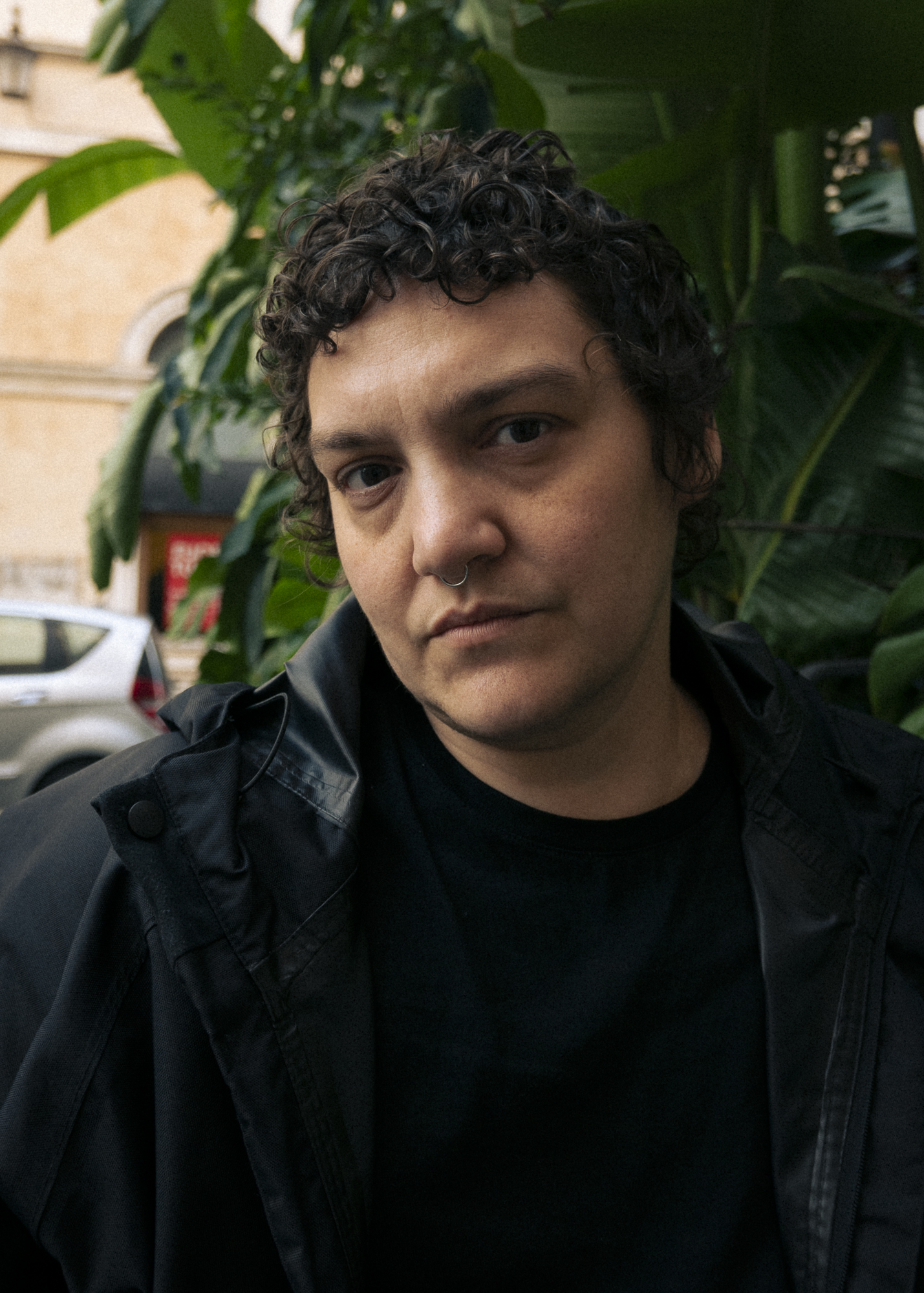 Portrait of Elle Perez. Perez is looking directly at the camera with their head tilted to the side. They are standing in front of what appears to be a wall of vibrant green plants with large leaves. To the left of the frame a silver car and a washed tan brick building are visible. Perez is dressed in a black t-shirt and raincoat and their curly hair is cropped short.