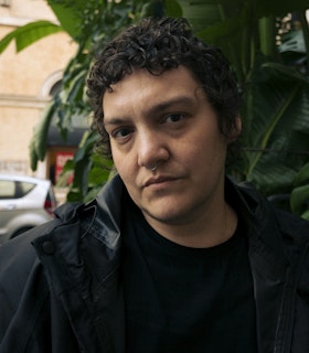 Portrait of Elle Perez. Perez is looking directly at the camera with their head tilted to the side. They are standing in front of what appears to be a wall of vibrant green plants with large leaves. To the left of the frame a silver car and a washed tan brick building are visible. Perez is dressed in a black t-shirt and raincoat and their curly hair is cropped short.