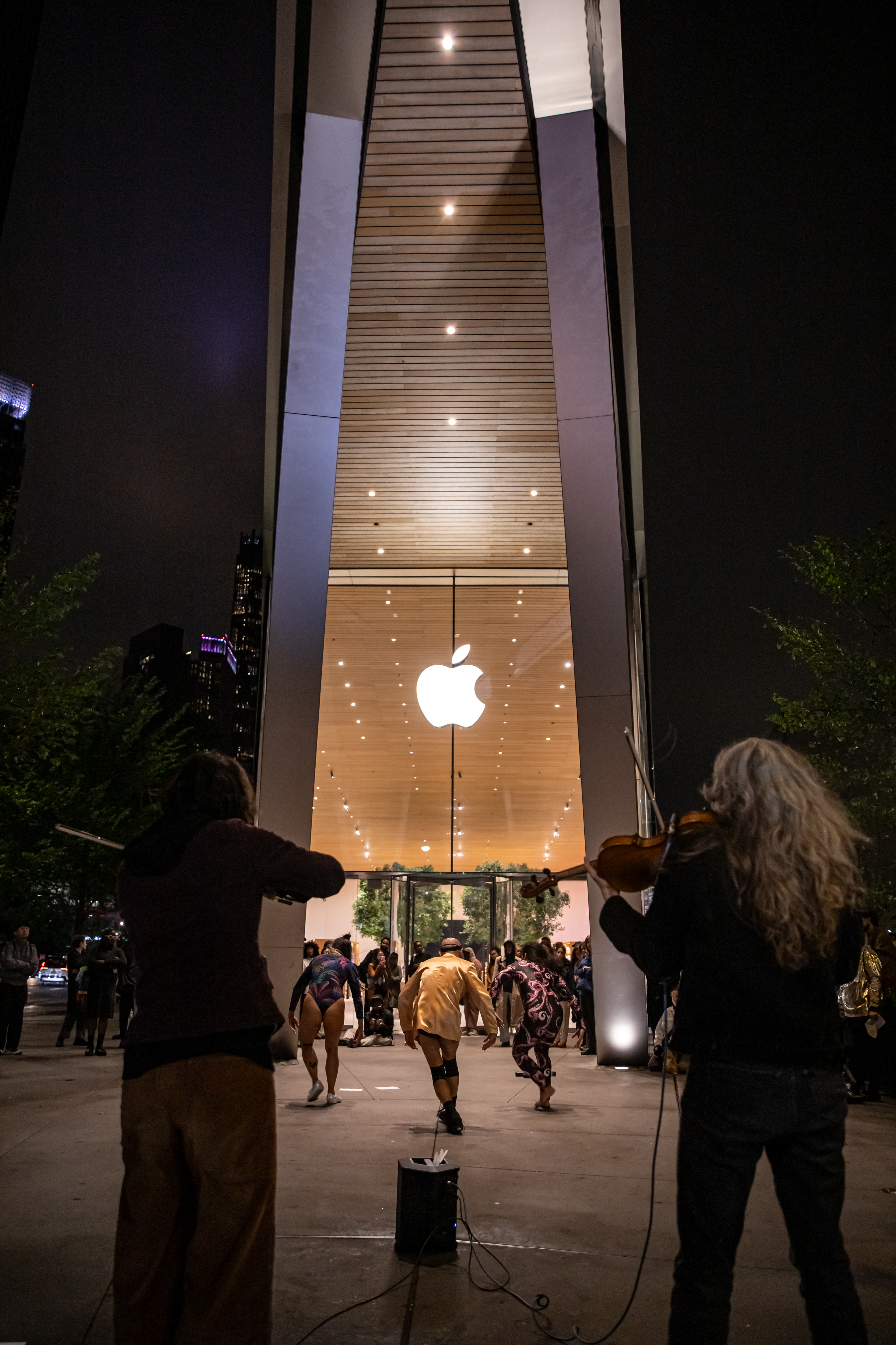 Two musicians play amplified violins outside in the  evening, facing away from the camera and towards  three dancers, who are in front of a brightly lit Apple  Store in downtown Brooklyn. The store has a tall glass  facade, which overwhelms the center of the image.  Audience members watch the performance from  outside of the store and the street.