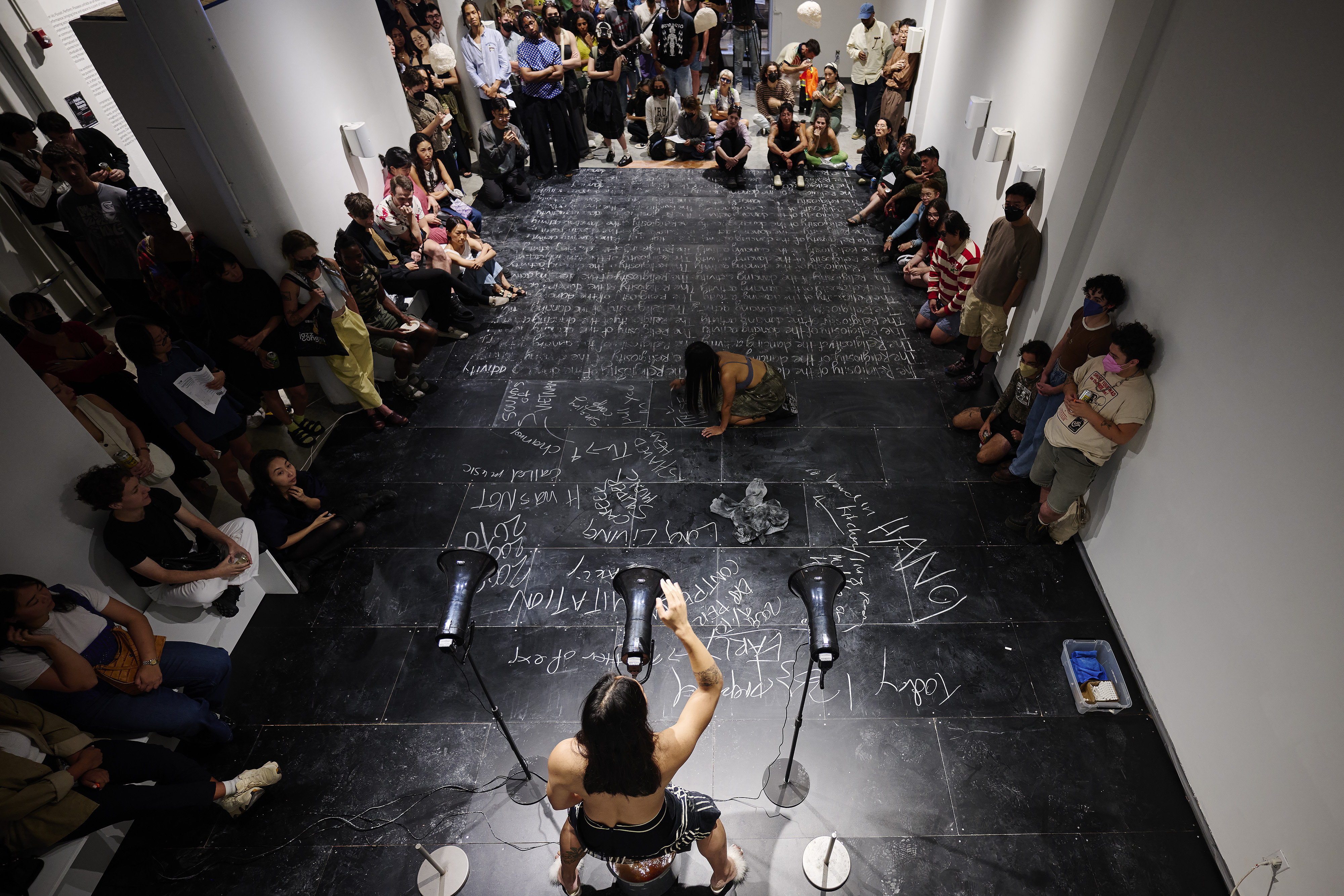 Facing away from the camera, Anh Vo holds their right  hand up in front of three black megaphones on stands.  In the center of the image, in front of Vo, Kristel Baldoz  is on the ground on all fours, writing with chalk  asymmetrically. Behind Kristel is additional writing in chalk,  which appears to be the same words written over  and over again. Audience members are seen throughout the sides and back of the room.