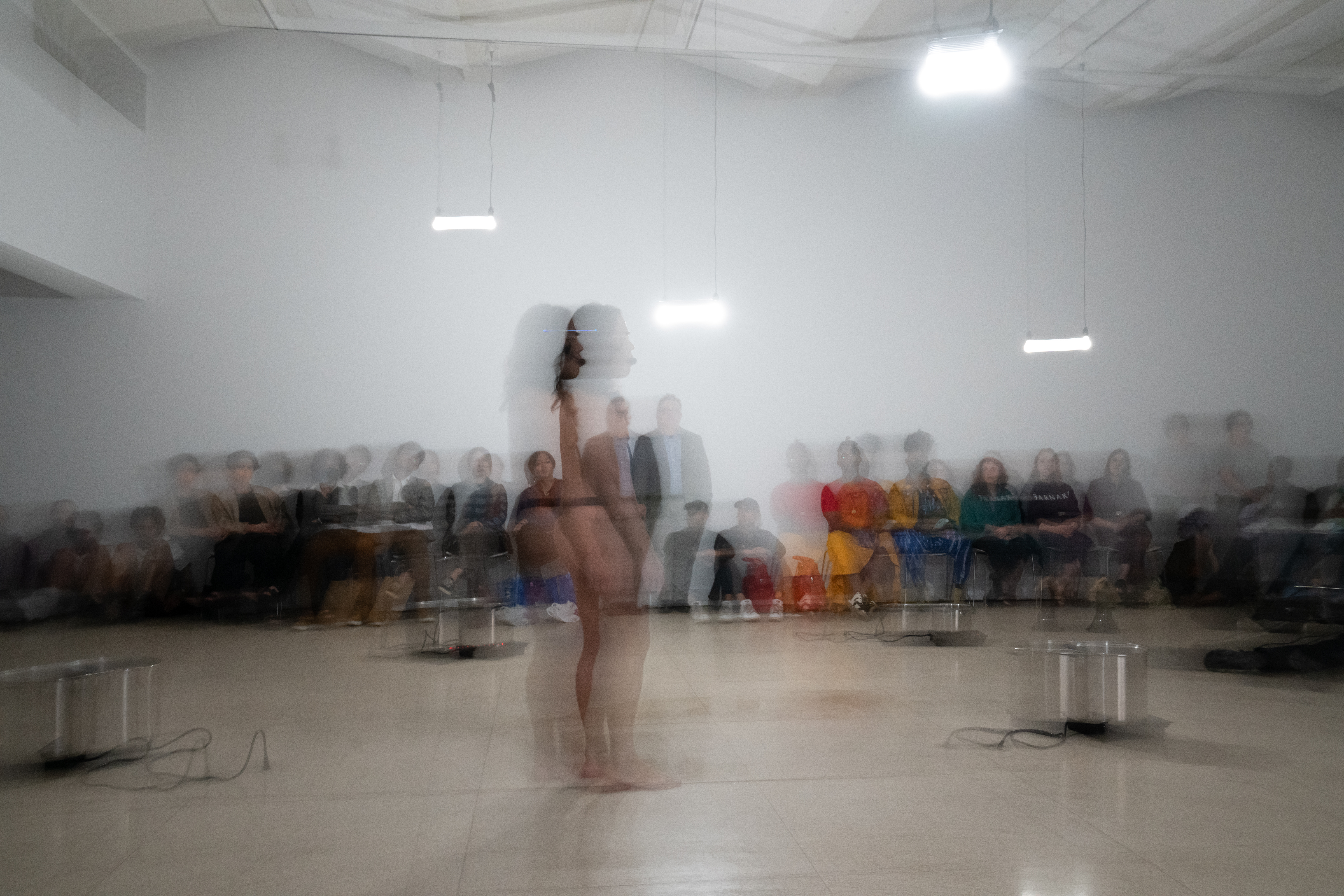 A long exposure shot of Anh Vo performing at The Kitchen.  They are in the center of the image, facing to the side,  encircled by four large plugged-in steam cookers. Anh Vo is  only wearing underwear. Audience members are seated  against the white wall. Bright white lights hang from the  ceiling grid.