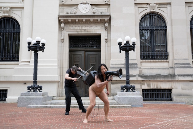 Anh Vo in nude underwear and an amplifier backpack,  shouting into a black megaphone on the sidewalk in front  of the American Numismatic Society building. Their legs are  spread out, with the wire from the megaphone falling behind  and in between. To the left and behind of Anh Vo is Ethan  Philbrick, wearing all black and playing the cello.