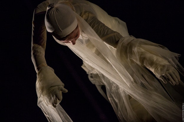 Maggie Cloud looking down as she performs. Her hands are facing out and she is  wrapped in gauzy fabric. The angle of this photograph makes Maggie appear to be  floating. The background is pitch black.