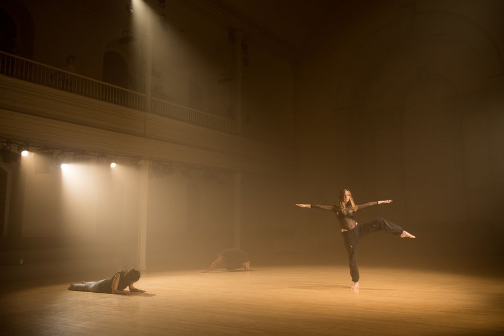 Three dancers performing in a dimly lit, foggy room. On the right hand side, one  dancer is in a pirouette-like position, extending both of their hands and one leg to  the sides. In the center and to the left are two more dancers, moving on the ground.