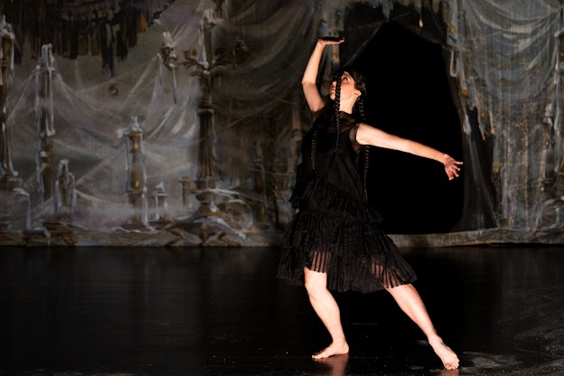 Maggie Cloud performing. She is looking at her hand above her head as she  extends her other arm to the side and points the toes on one of her feet, as if  she is about to leap. Cloud is wearing a black dress. Behind her is a painted backdrop with melted candles on brown candle holders, white curtains that look  almost like webs, and a black chandelier that is matching her dress.