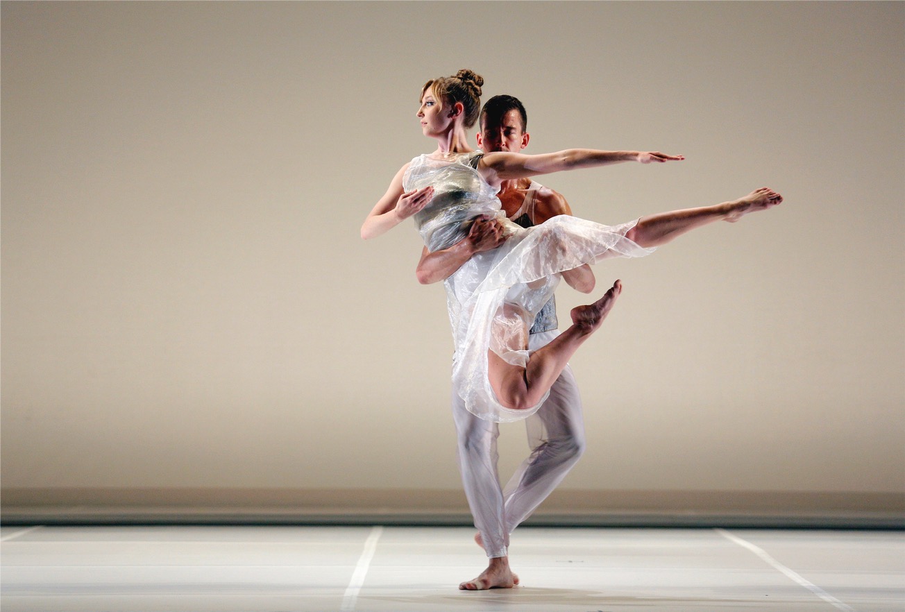 Dylan Crossman steps one foot in front of the other as he holds Maggie Cloud in  the air in front of a beige wall. Cloud's back is towards him and she looks to the  side while pointing her arms and feet. Both are wearing white semi-see through  garments with blue accents.