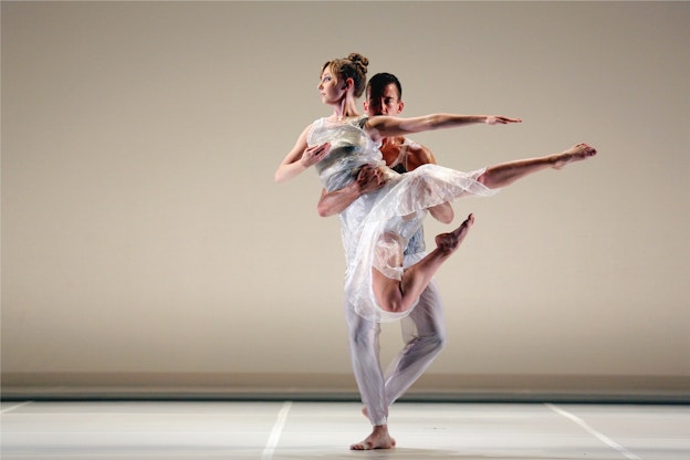 Dylan Crossman steps one foot in front of the other as he holds Maggie Cloud in  the air in front of a beige wall. Cloud's back is towards him and she looks to the  side while pointing her arms and feet. Both are wearing white semi-see through  garments with blue accents.