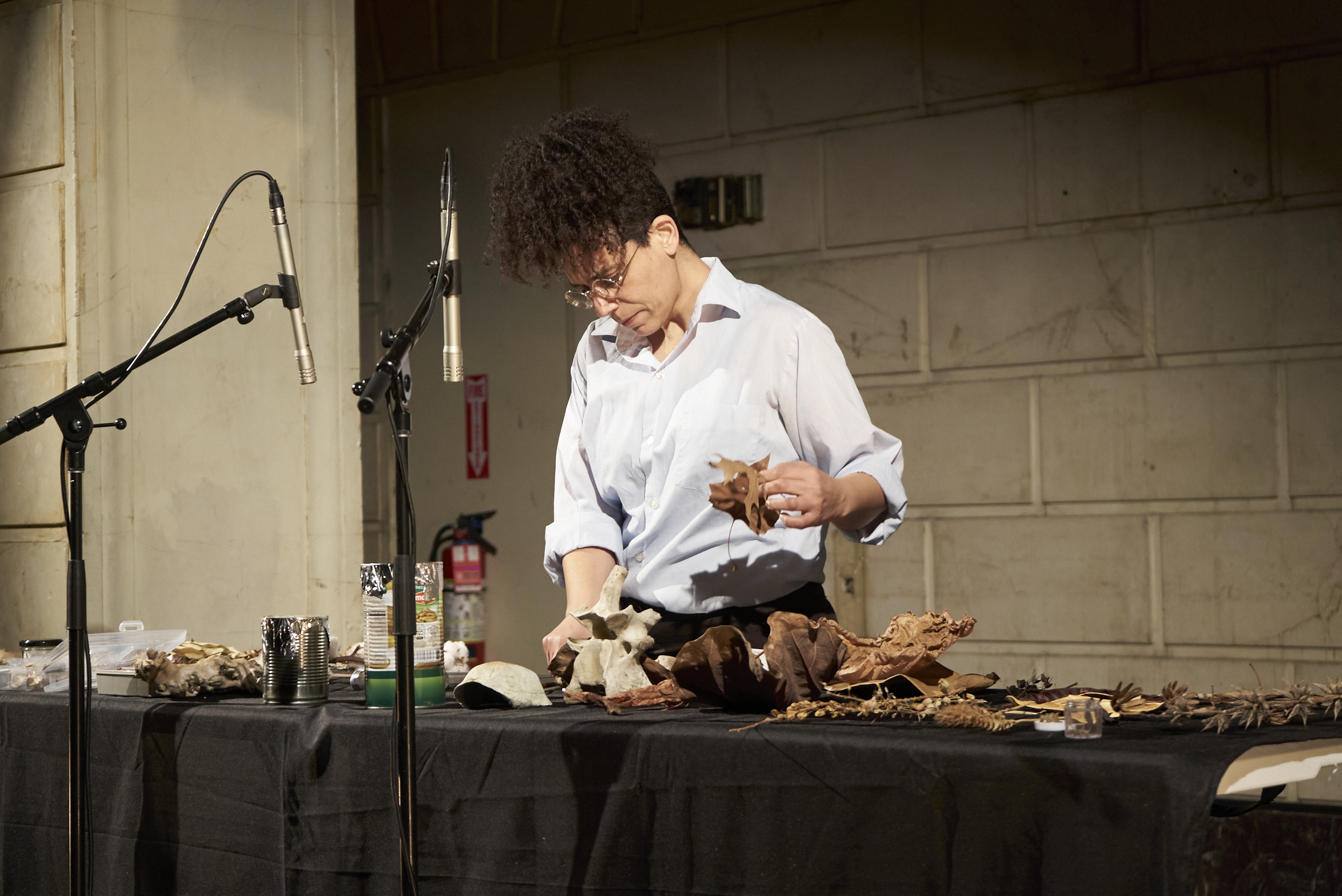 Maayan Tsadka mid-performance. She is wearing a white collared shirt, glasses, and her hair pulled up.  She is standing over a long table covered in a black tablecloth. In Tsadka’s hand she holds a leaf and on  the table is an assortment of different objects being used in the performance, including dried leaves and  plants, bones, shells, and metal cans covered in foil. In front of her and the table are two microphones on  stands pointed downwards towards the objects table.